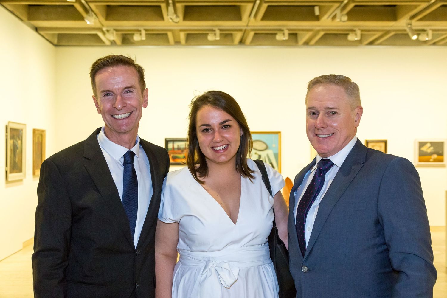 Two men in suits and a smiling woman in a white dress pose together inside a bright art gallery with framed paintings on the walls.