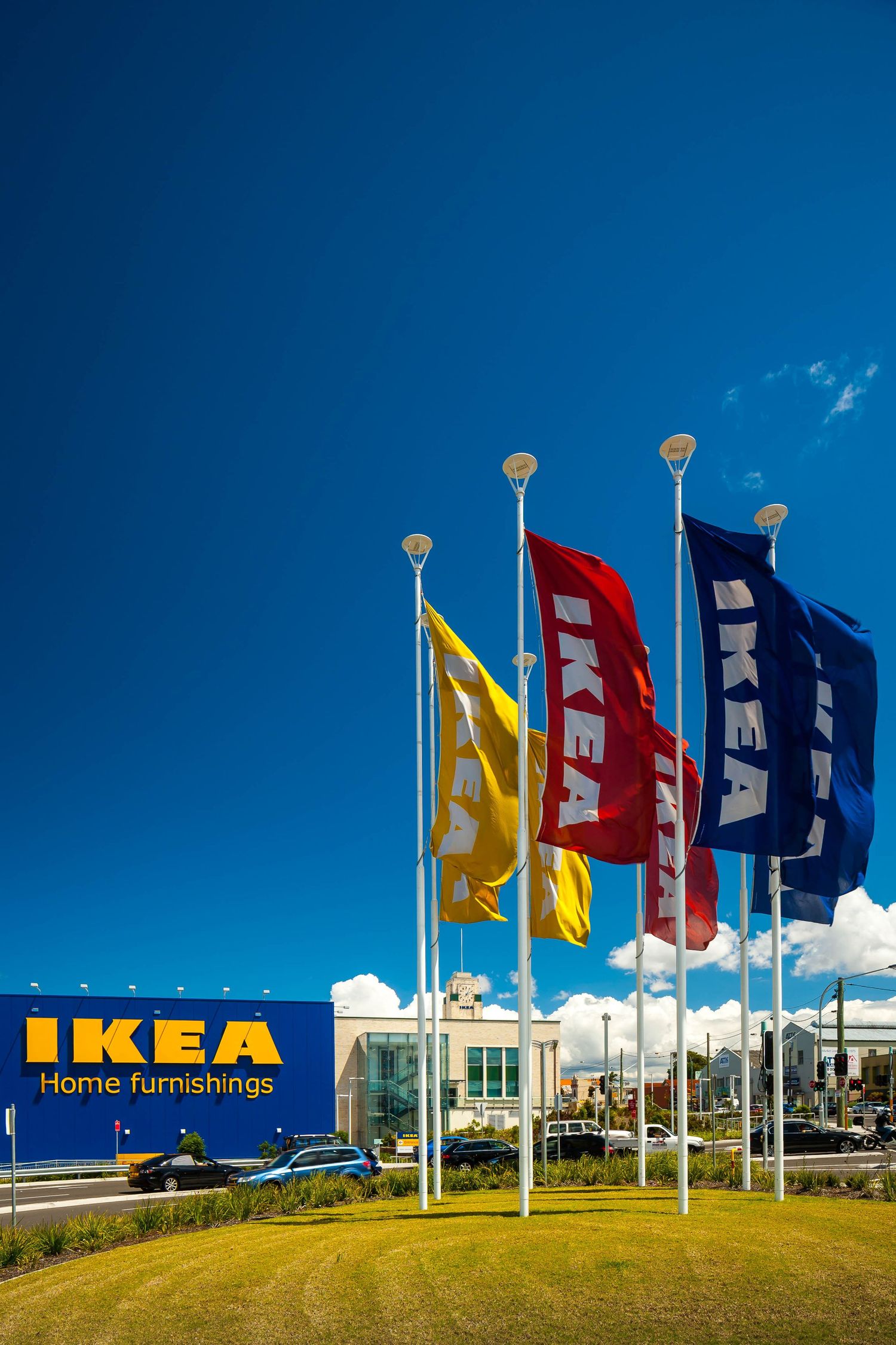 Blue, red and yellow IKEA flags fly on tall poles in front of an IKEA home furnishings store under a bright blue sky.