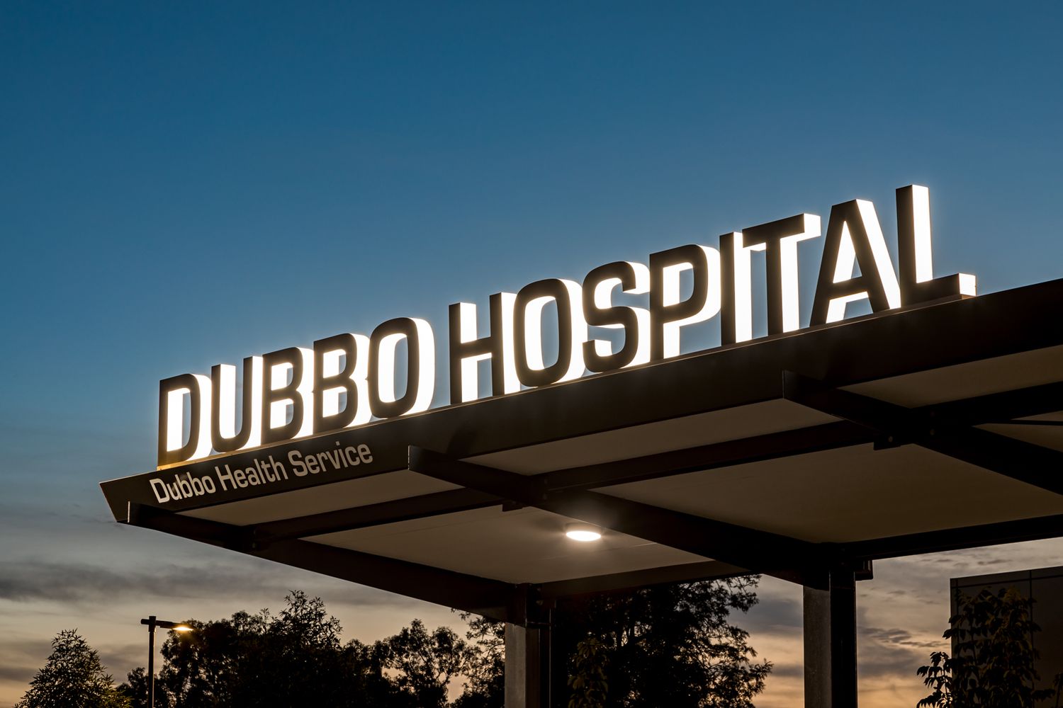Illuminated Dubbo Hospital sign on a canopy entrance at dusk, marked Dubbo Health Service, under a deep blue evening sky.