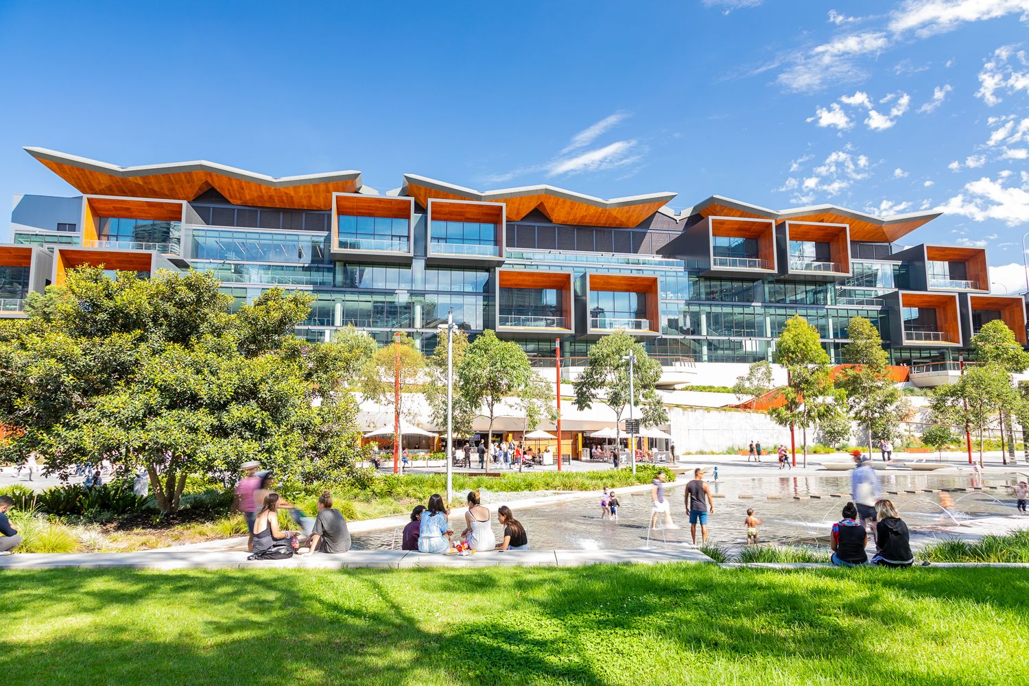 Modern multi-story building with orange timber-style roof accents and glass facade, with people relaxing on a sunny lawn and plaza.