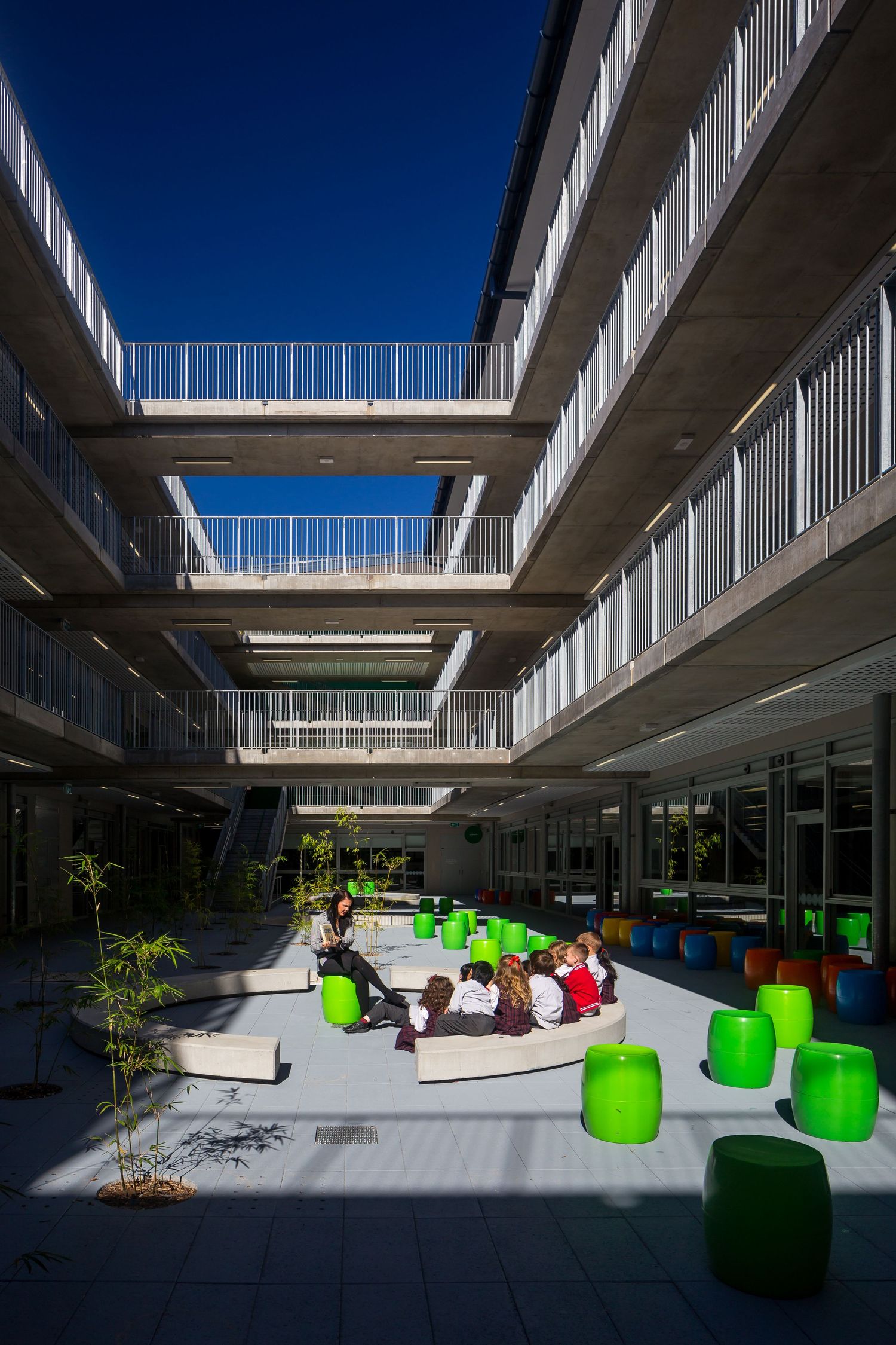 Sunlit school courtyard with multi-level concrete balconies, green stools and children gathered around a seated teacher.