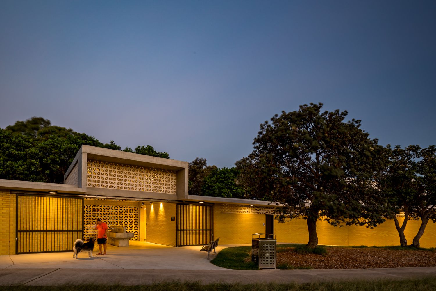 Mid-century modern yellow building at dusk with warm glowing lights, a breeze block screen, and a person walking a dog outside.