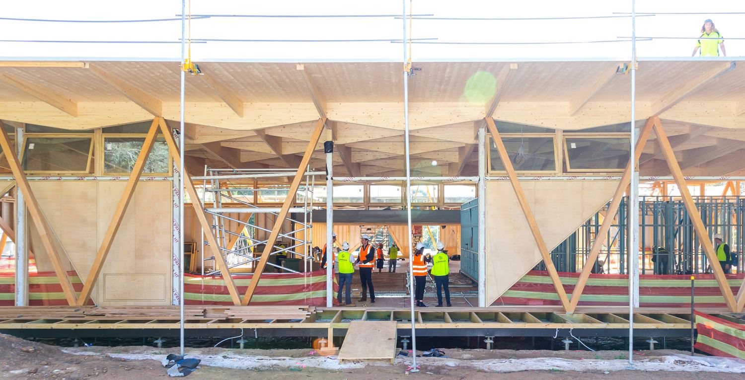 Construction workers in hi-vis vests stand inside a partially built timber-framed building with exposed diagonal wooden beams.