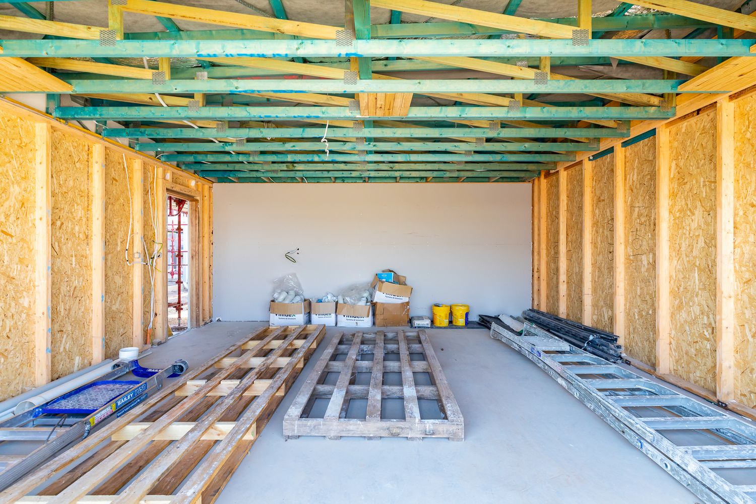 Unfinished interior with exposed timber roof trusses, OSB wall sheeting, stacked building supplies and wooden pallets on a concrete floor.