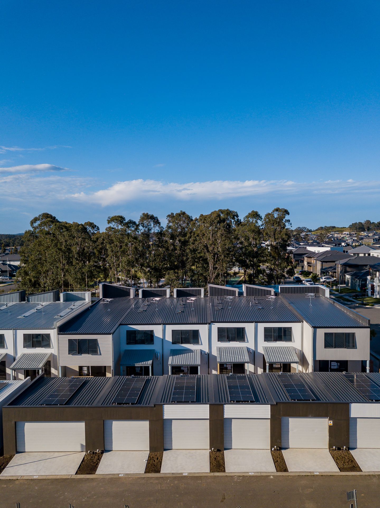 Aerial view of modern townhouses with solar panels on the roofs and garages in a new estate beside a line of gum trees.