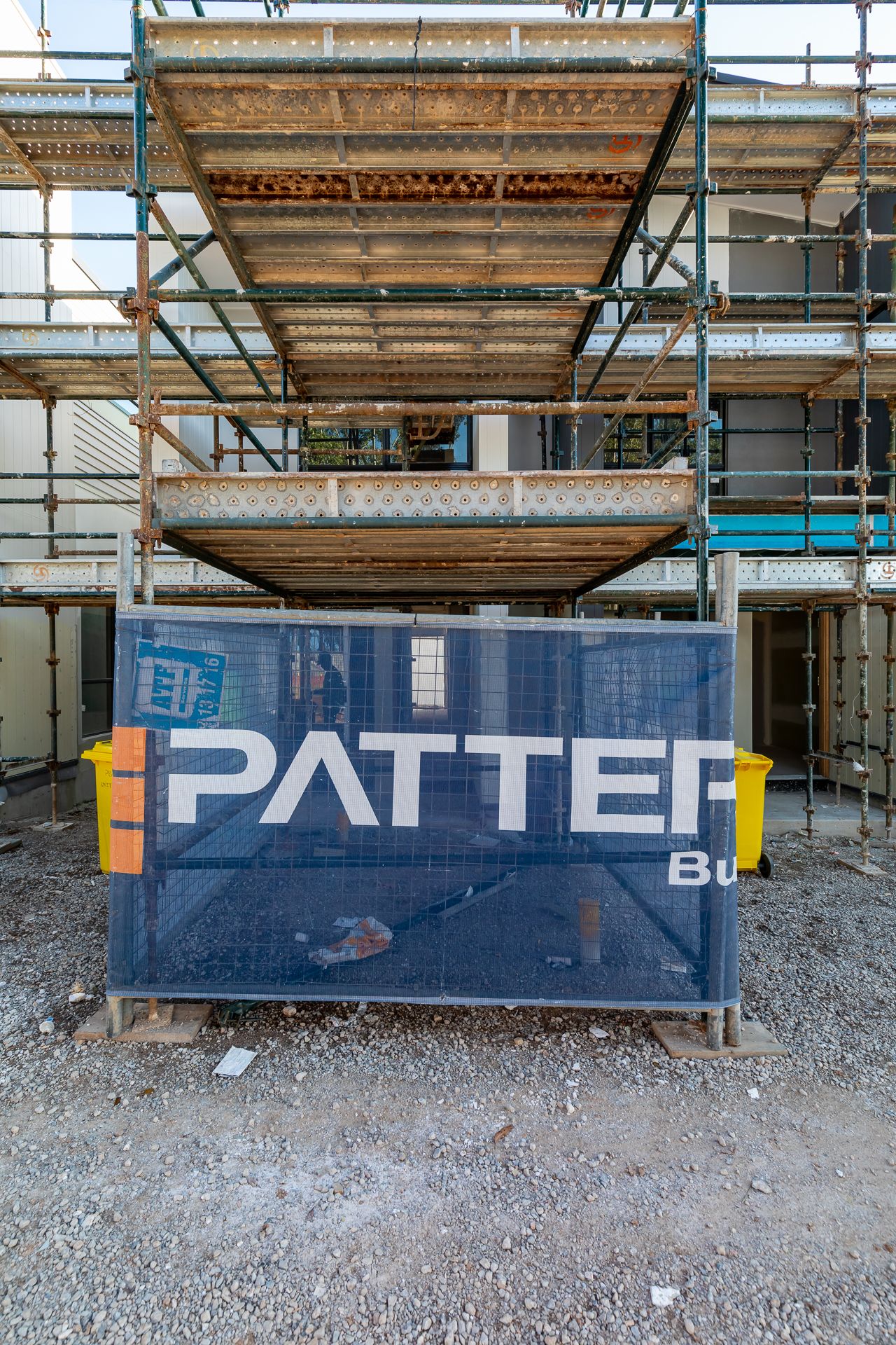 Construction site scaffolding behind a blue safety fence banner showing a building company name, with gravel ground in the foreground.