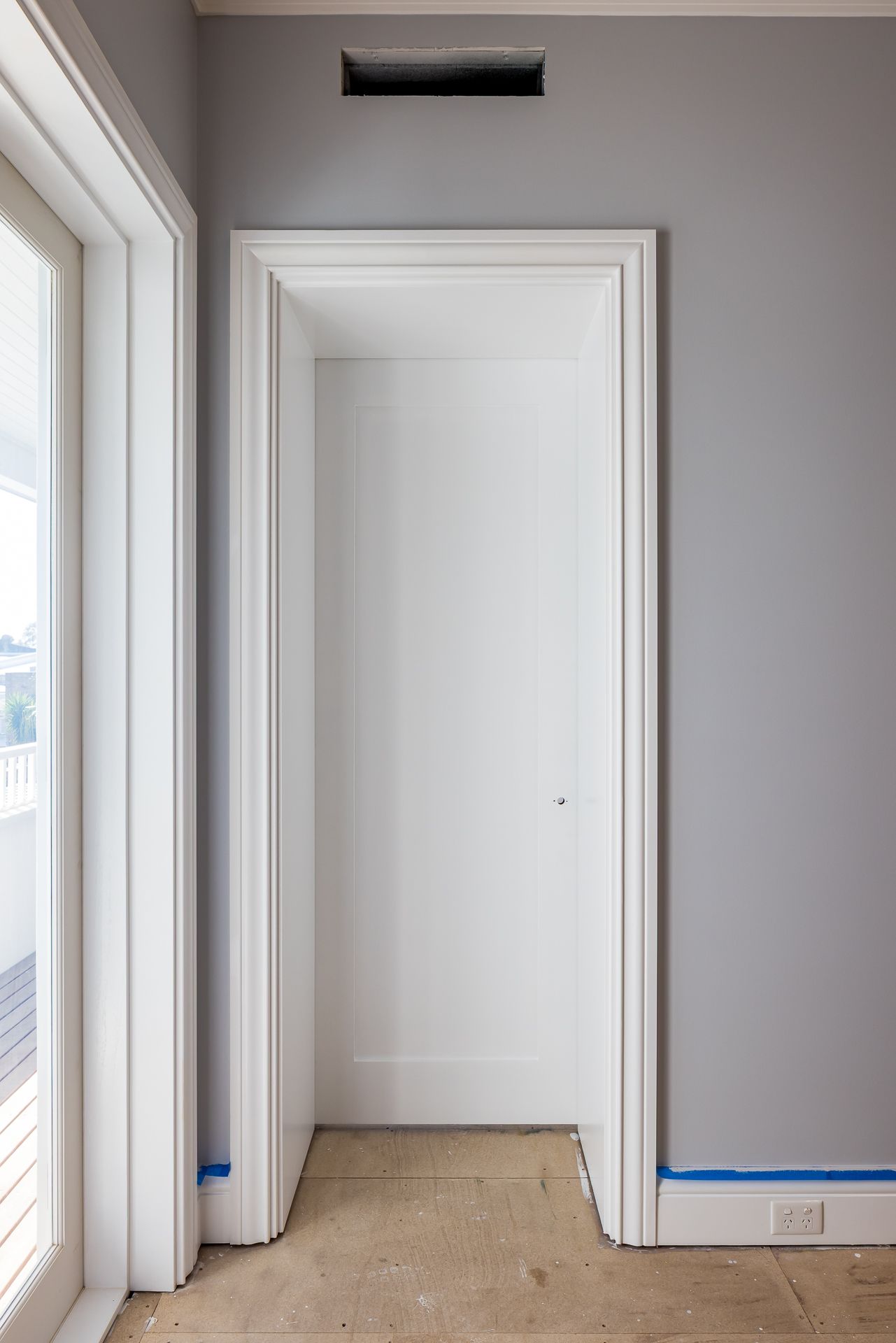 Unfinished interior with a white paneled door in a deep casing, grey walls, blue masking tape and bare subfloor beside a glass door.