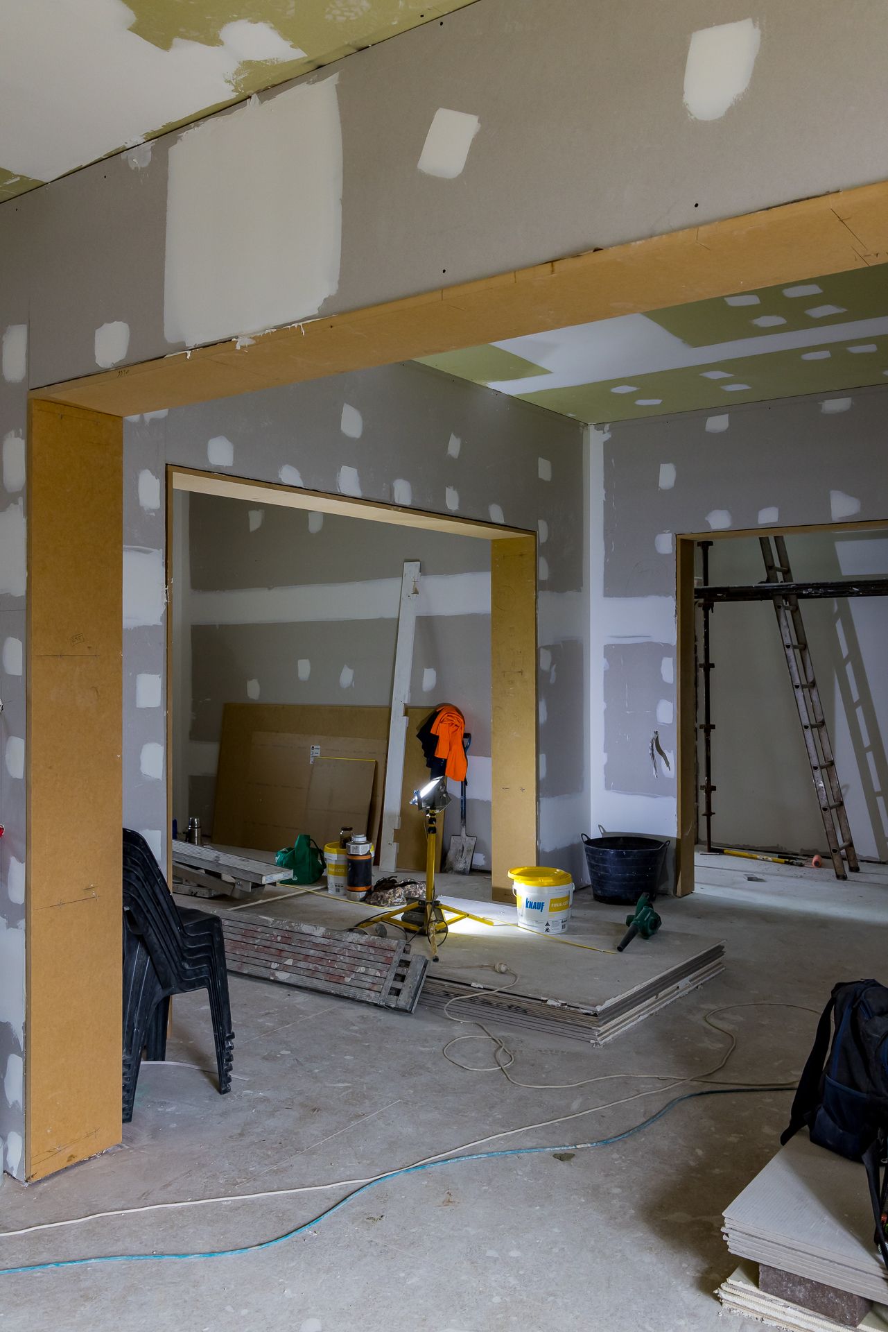 Home interior under renovation with taped drywall, wooden door frames, a ladder, paint buckets and tools on a dusty concrete floor.