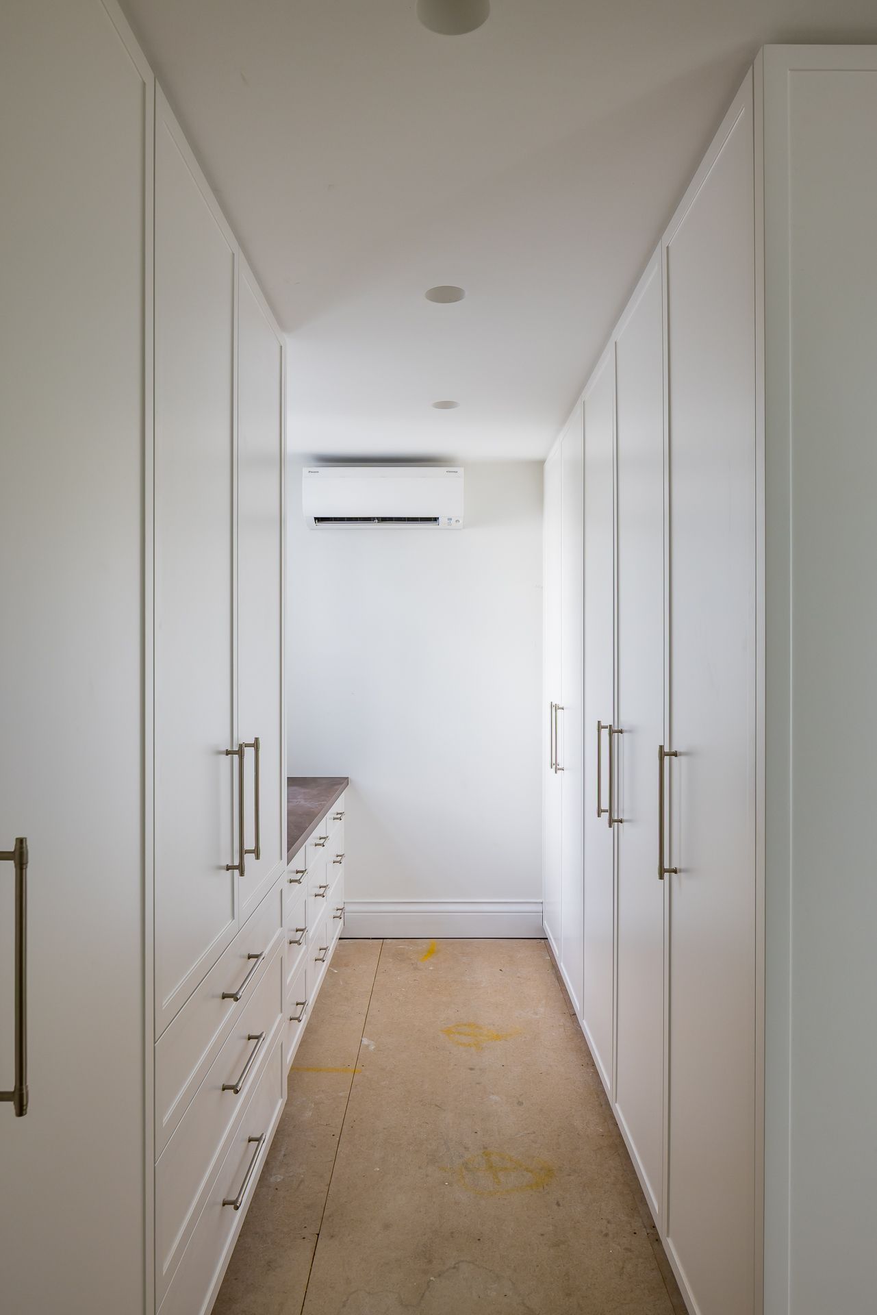 Narrow walk-in wardrobe with white floor-to-ceiling cabinets, brushed metal handles, drawers and a wall-mounted air conditioner.