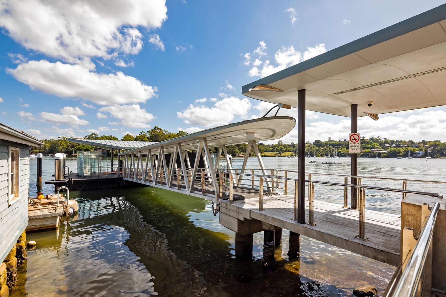 Harbour ferry wharf with canopy roof and steel truss gangway over calm water, with moored boats and trees under a partly cloudy sky.