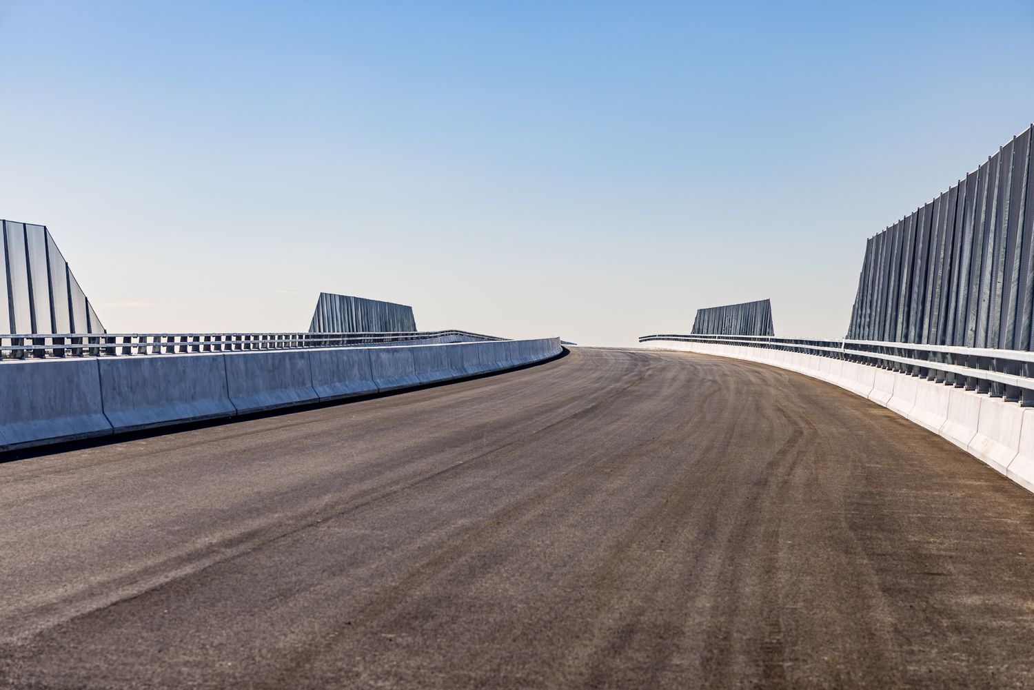 Empty newly constructed motorway bridge deck with smooth asphalt, concrete barriers and tall noise walls under a pale sky.