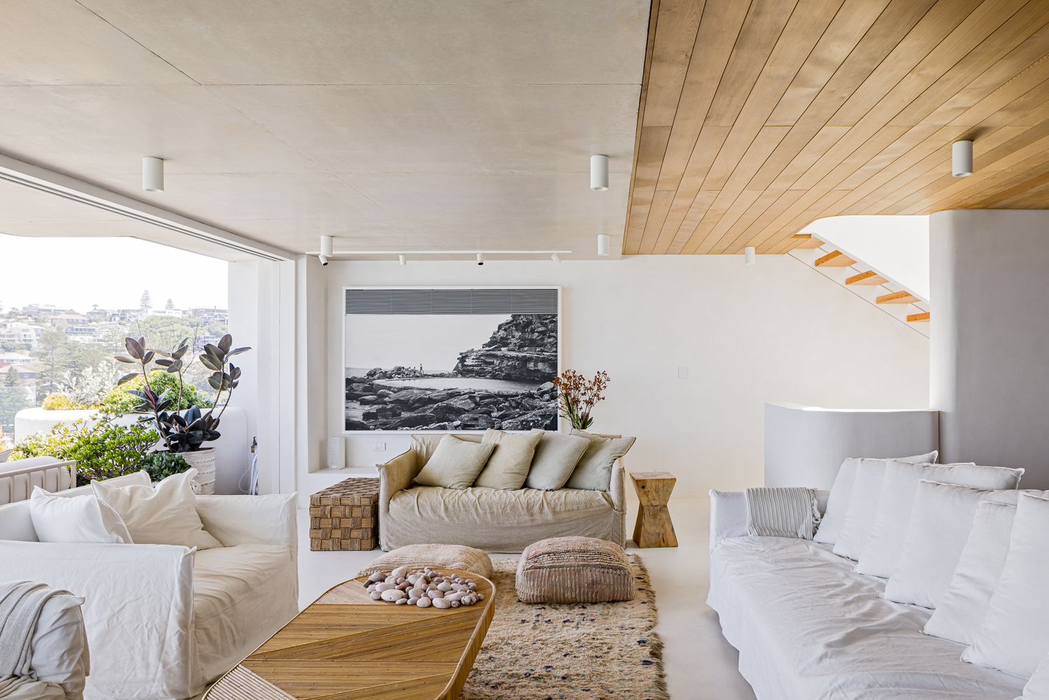 Bright lounge with white linen sofas, round timber coffee table, framed black-and-white coastal artwork and a timber-clad ceiling.
