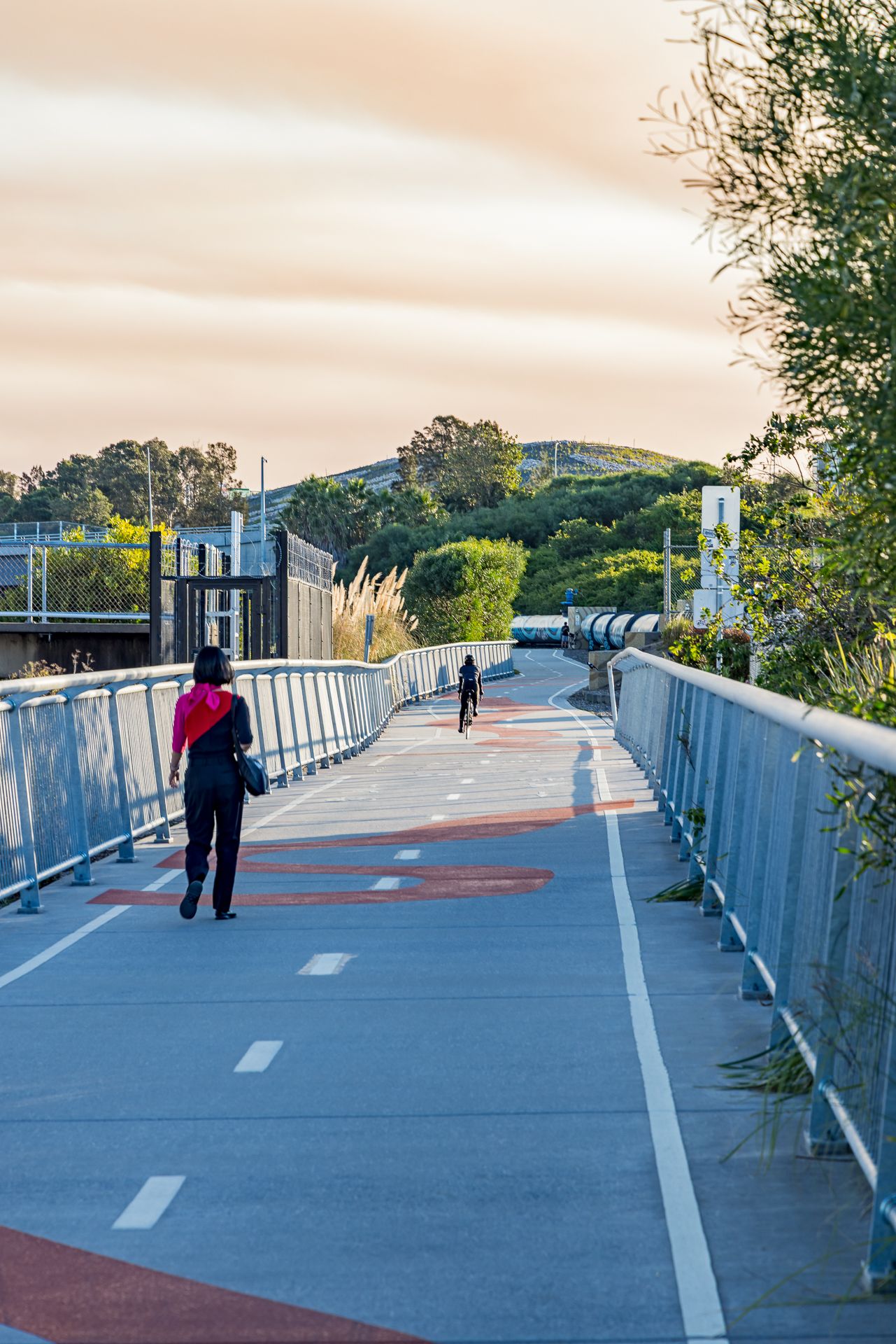 Pedestrian walking on a curved elevated bike path with red markings and metal railings at sunset.