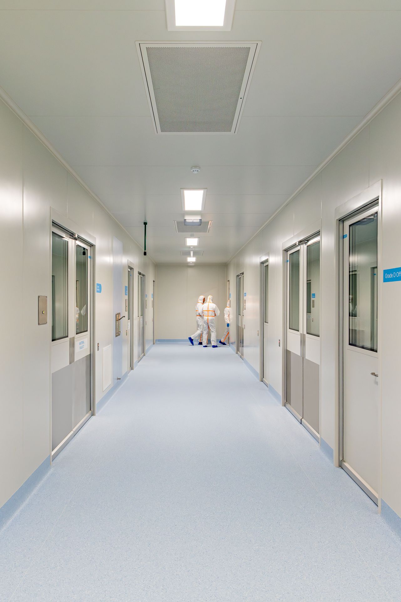 Long bright cleanroom corridor with glass-paneled doors and technicians in protective suits walking at the far end.