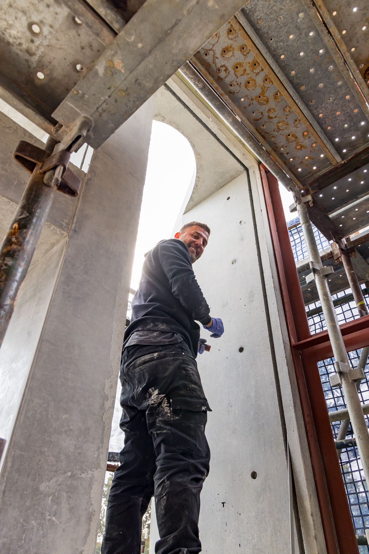 Smiling construction worker in dark work clothes and blue gloves stands among scaffolding beside a concrete wall and red window frame.