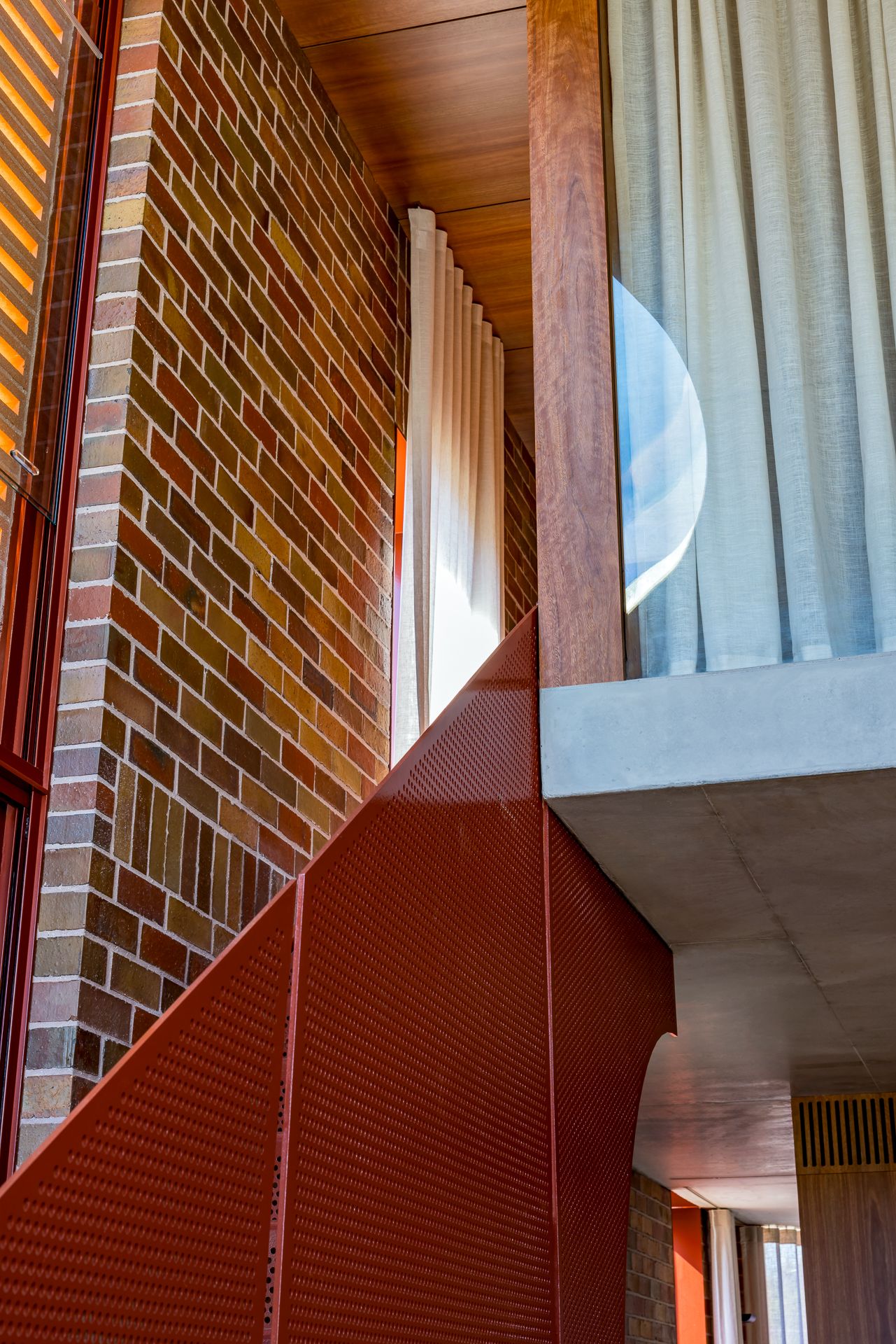 Modern interior with warm multicolored brick wall, wood ceiling, sheer curtains and a perforated rust-red metal staircase balustrade.