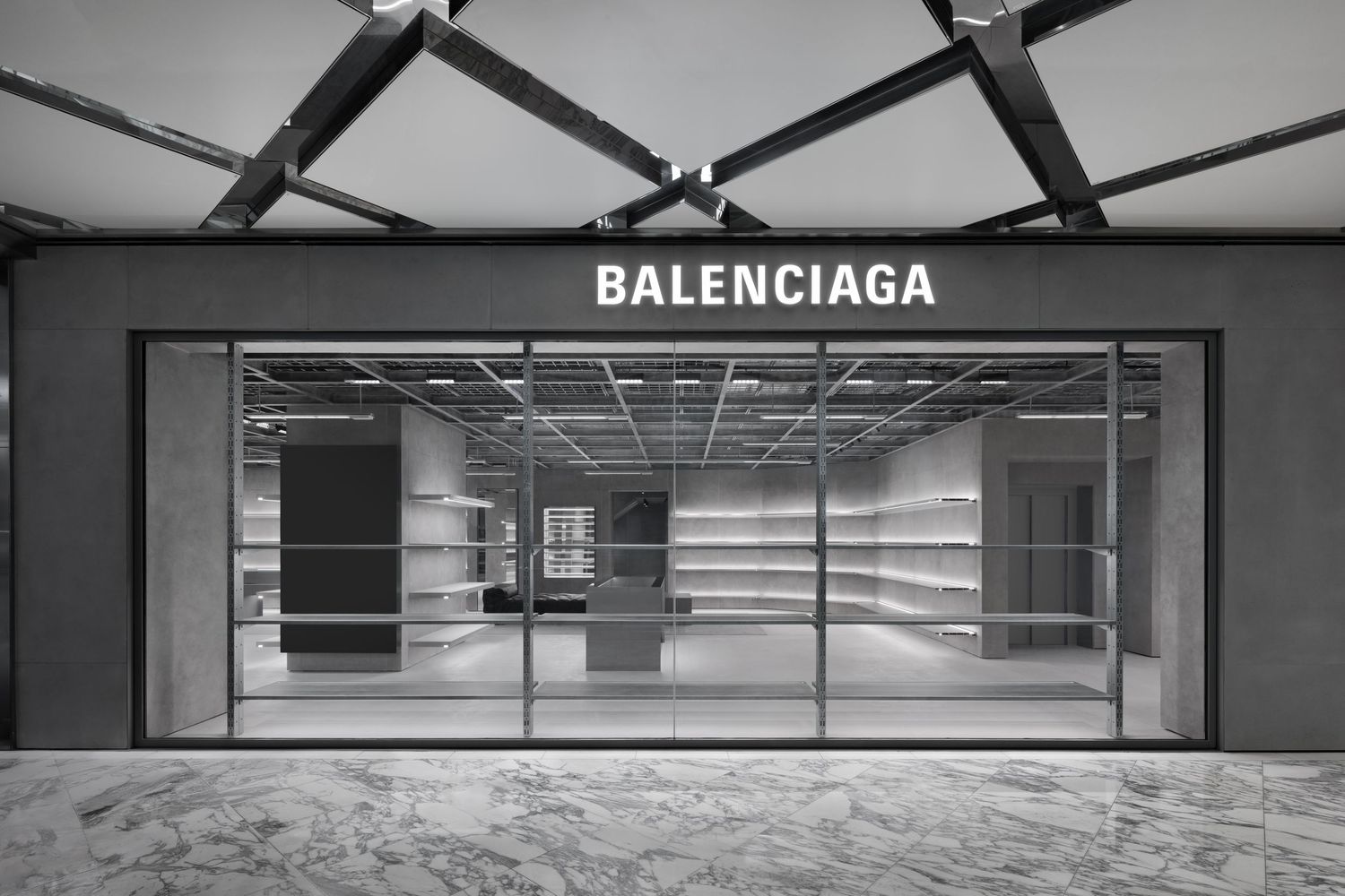 Balenciaga store inside a shopping mall with a black angular ceiling, glass frontage, marble flooring and minimalist white shelving.