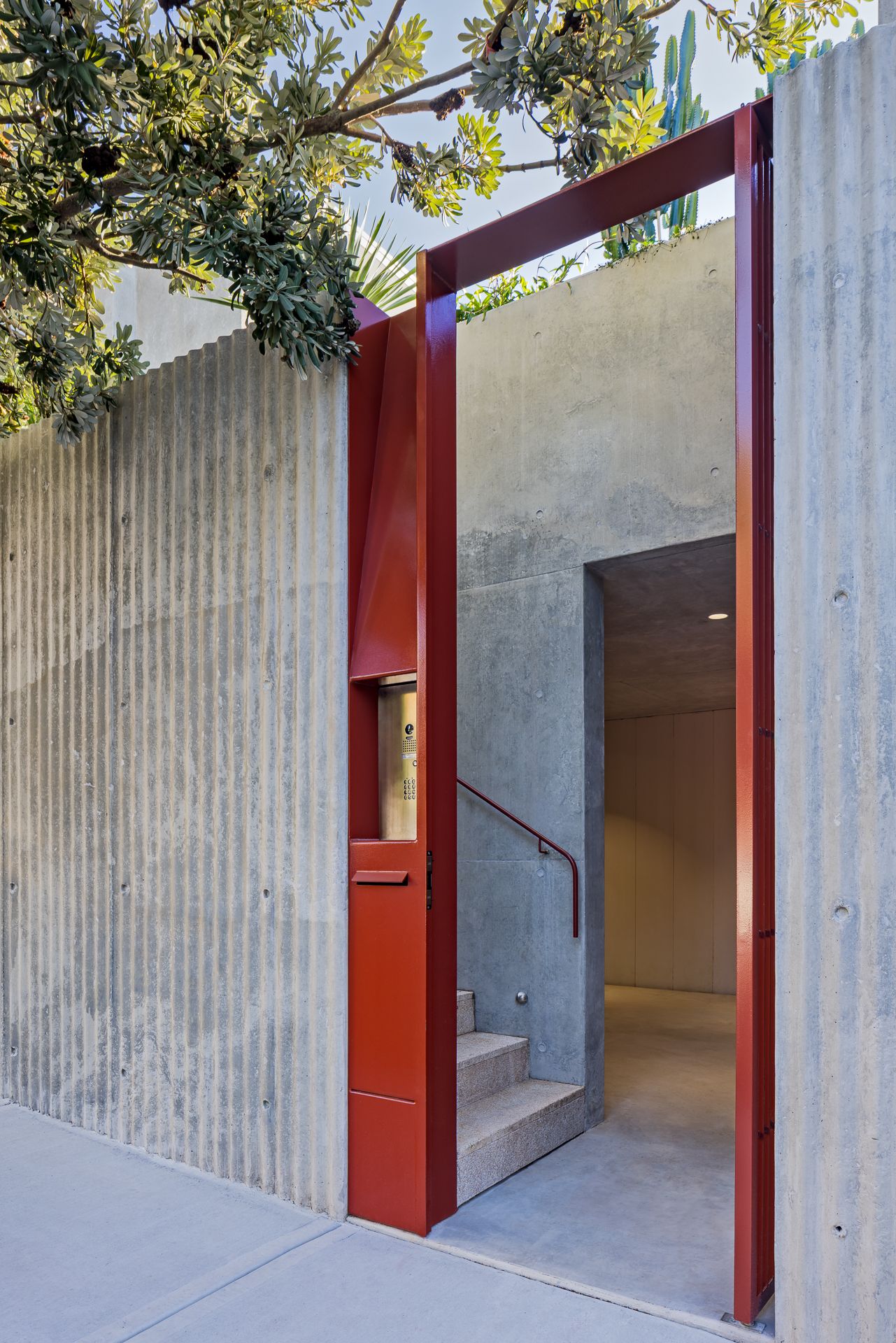 Red steel-framed entrance in a ribbed concrete wall leading to concrete steps, a handrail, and a softly lit interior.