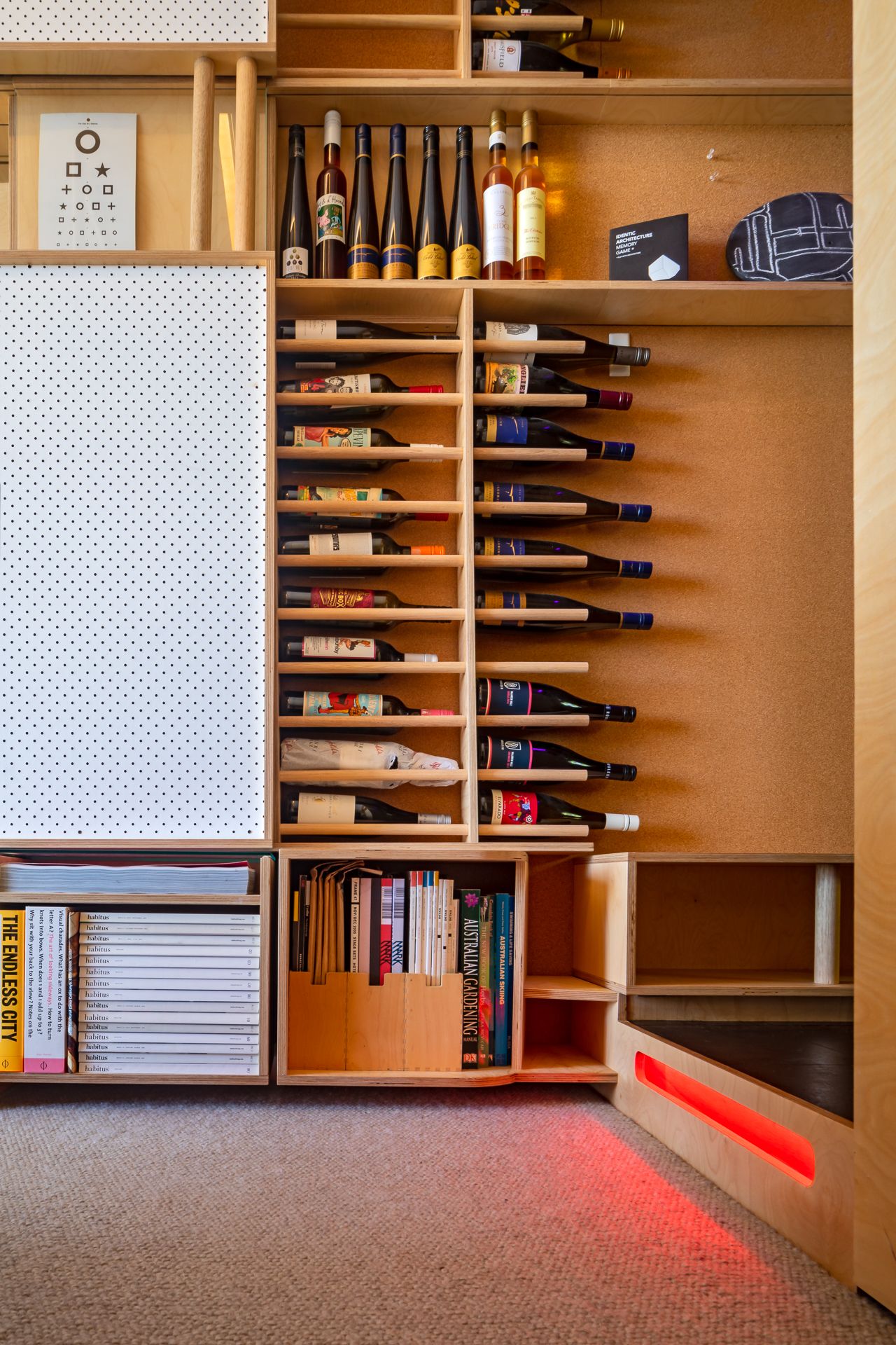 Built-in plywood shelving with slanted wine bottle storage, books and a glowing red step light above a gray carpet.