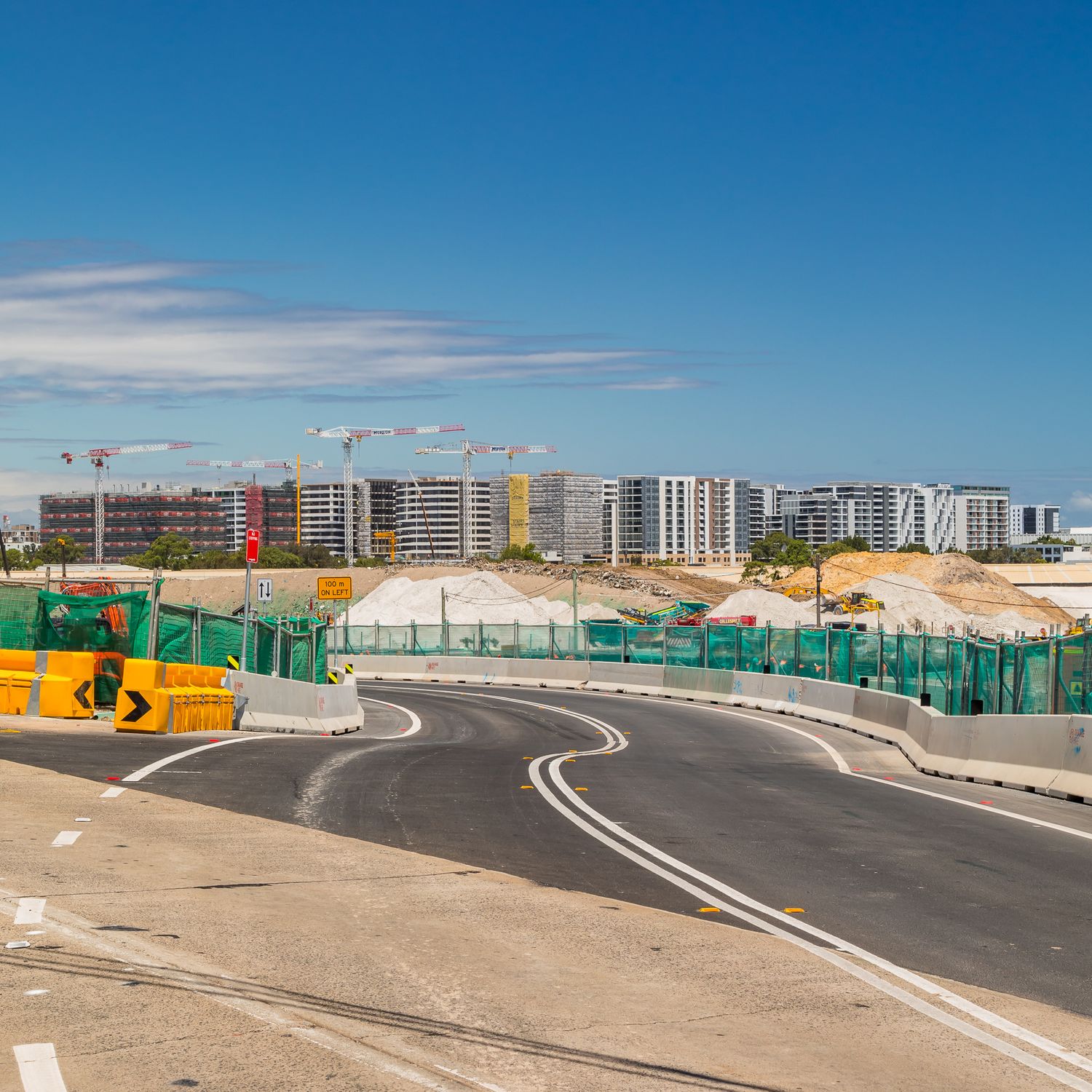 Newly built curved road with barriers and construction fencing leading toward a modern apartment development under construction.