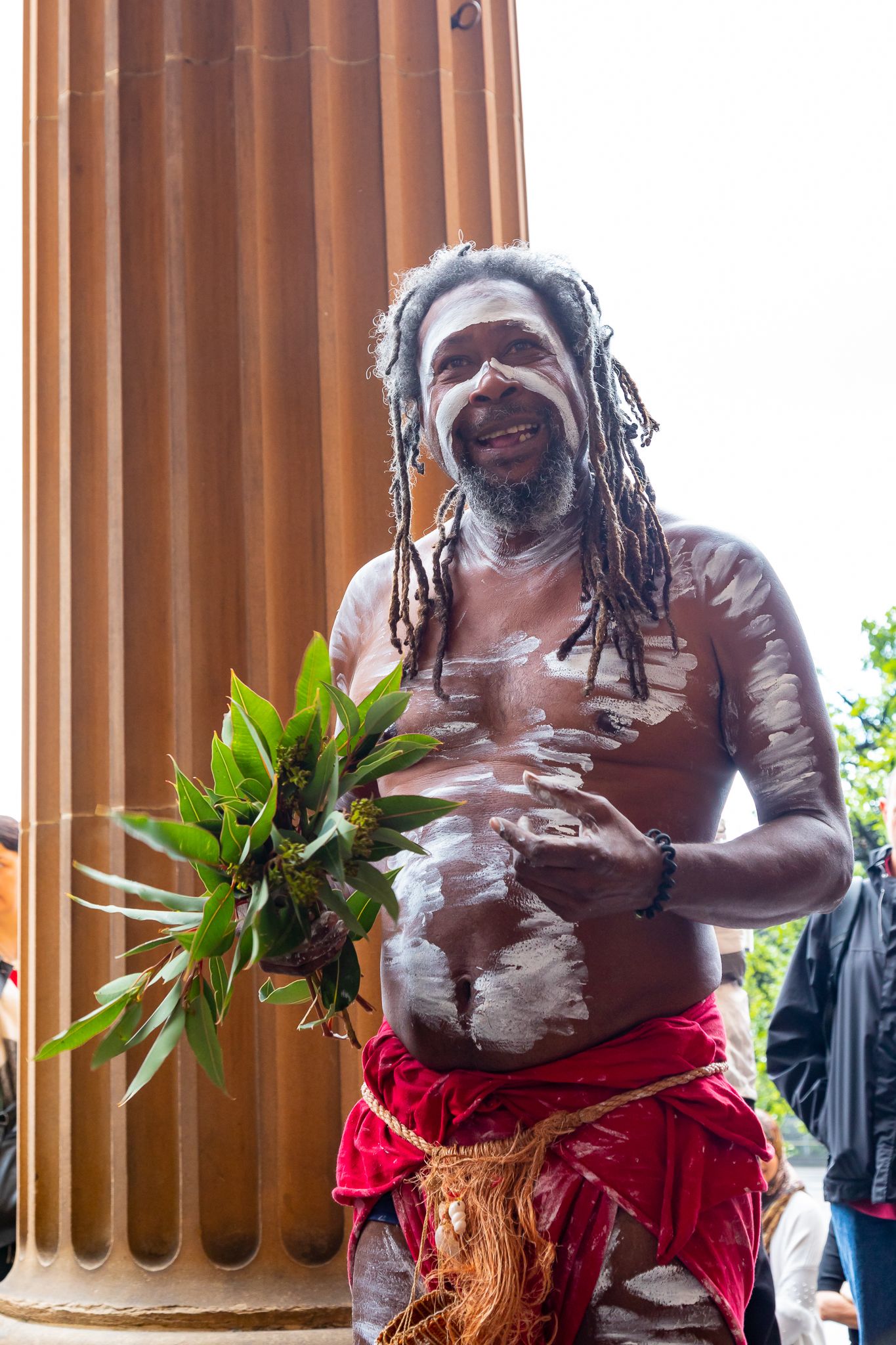 Smiling man with dreadlocks and white body paint holds a bunch of green eucalyptus leaves beside a large sandstone column.