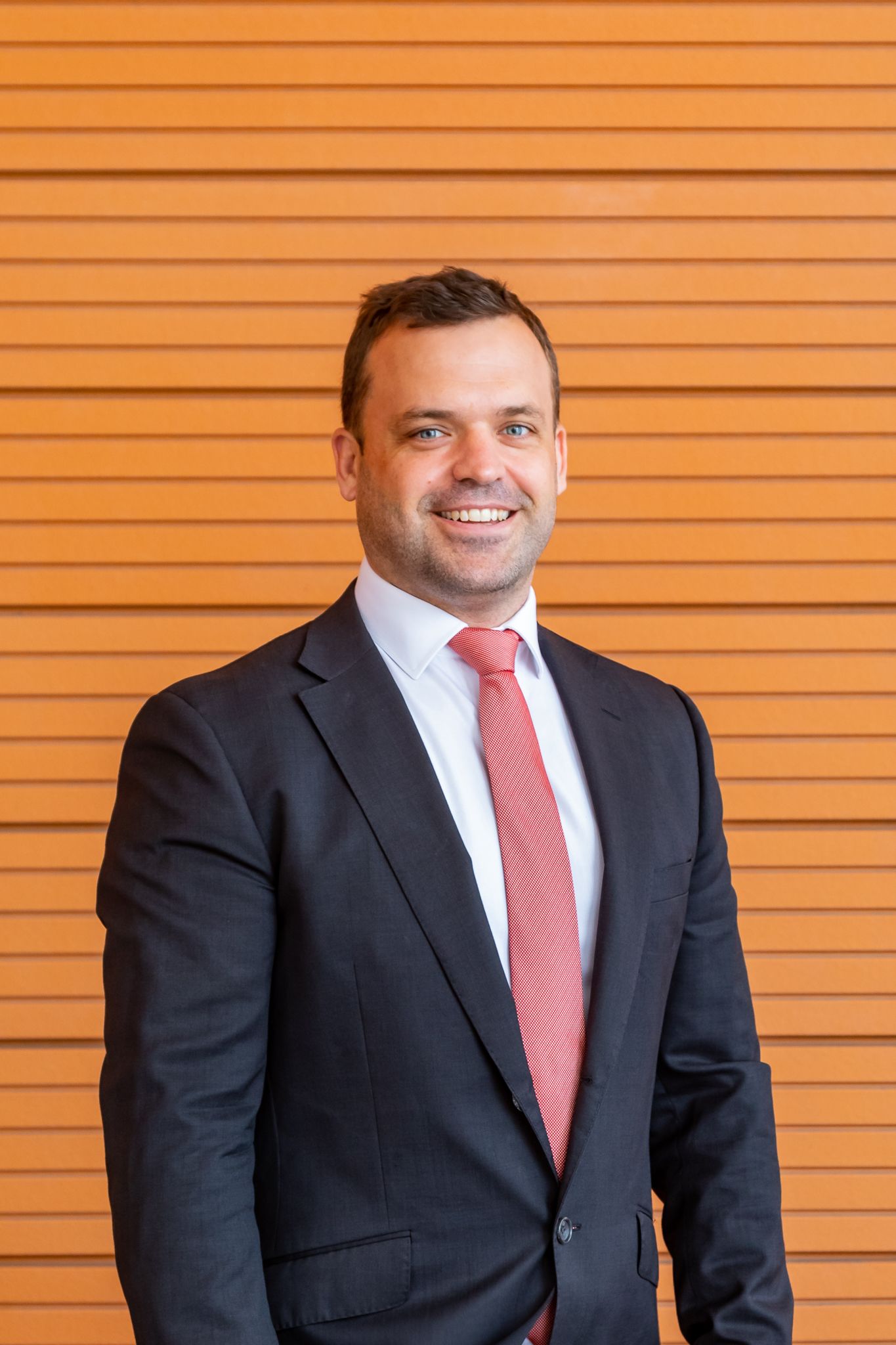 Smiling man in a dark suit, white shirt and coral tie stands in front of a horizontal wood-slat wall in a professional headshot.