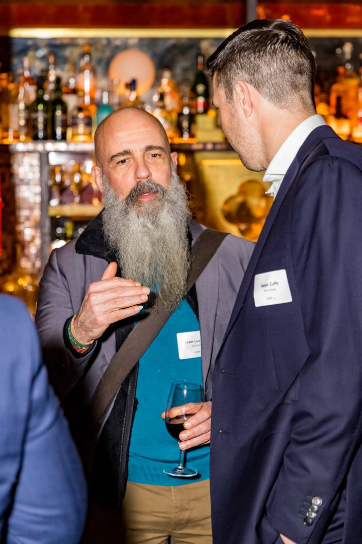 Bearded man gestures while talking to another guest at a networking event, both wearing name tags and holding wine glasses.