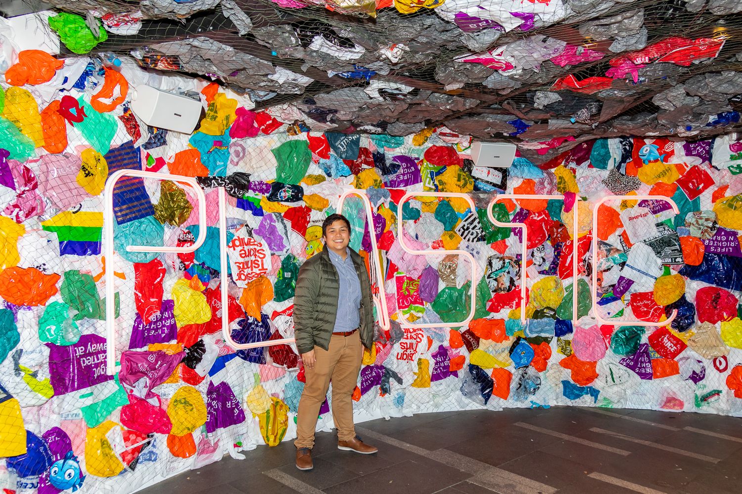 Man in an olive jacket stands inside a colorful art installation covered in bright pink, orange, green and purple shapes.