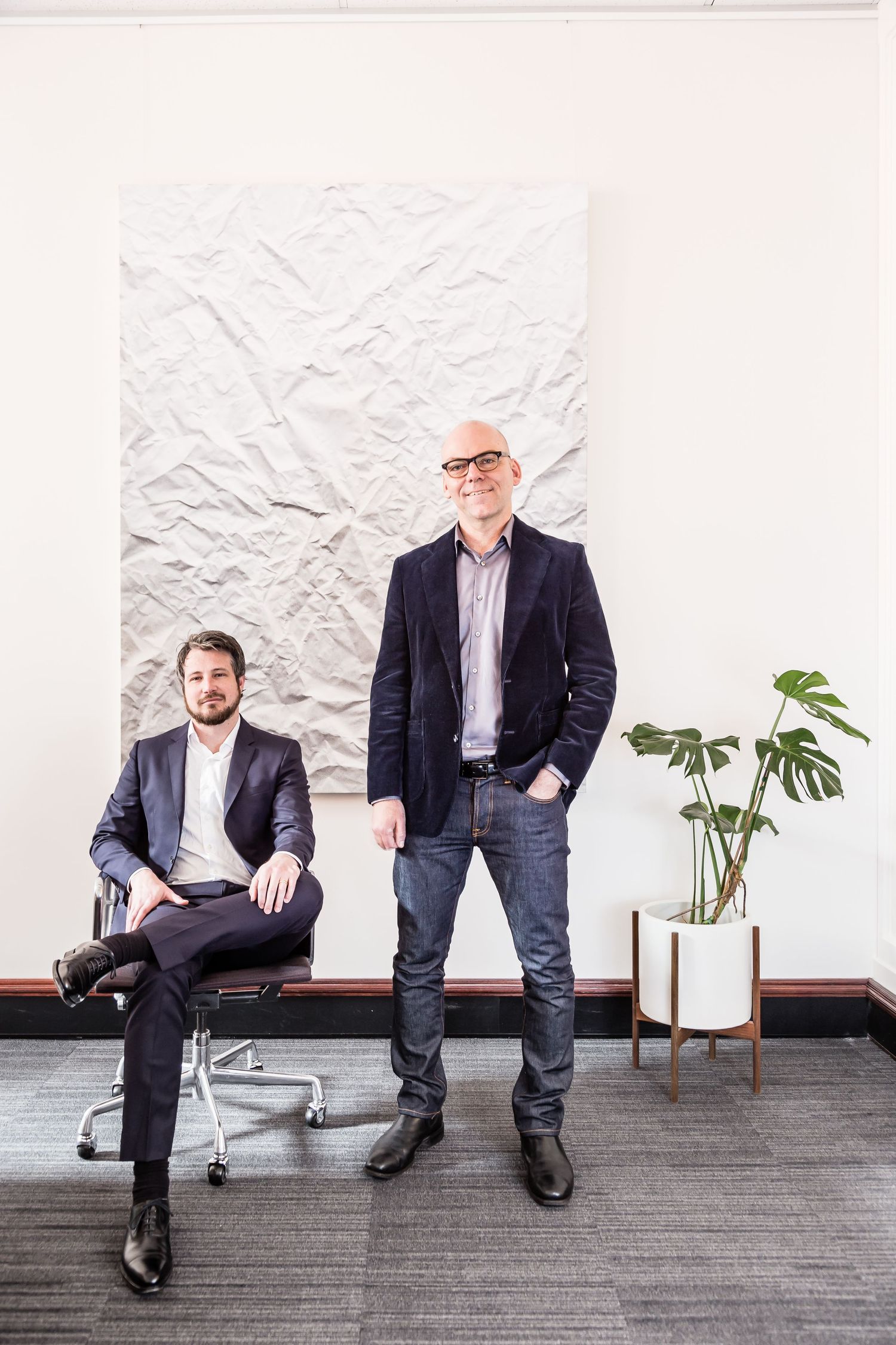Two men in blazers pose in a modern office, one seated on a chair and one standing beside a potted plant and a crumpled paper artwork.