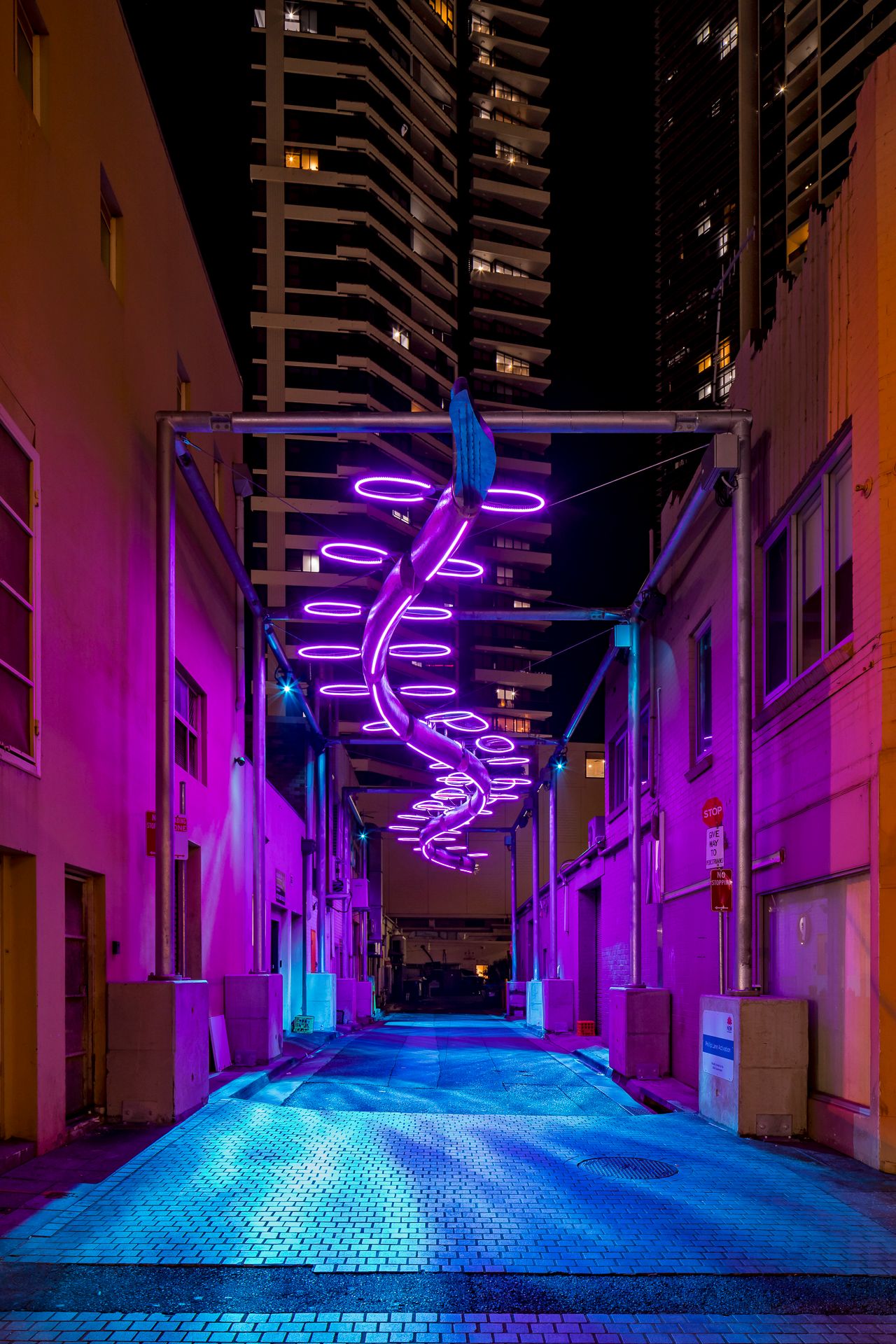 Empty urban alley at night lit in pink and purple by a spiral neon light sculpture, with a high-rise tower glowing in the background.