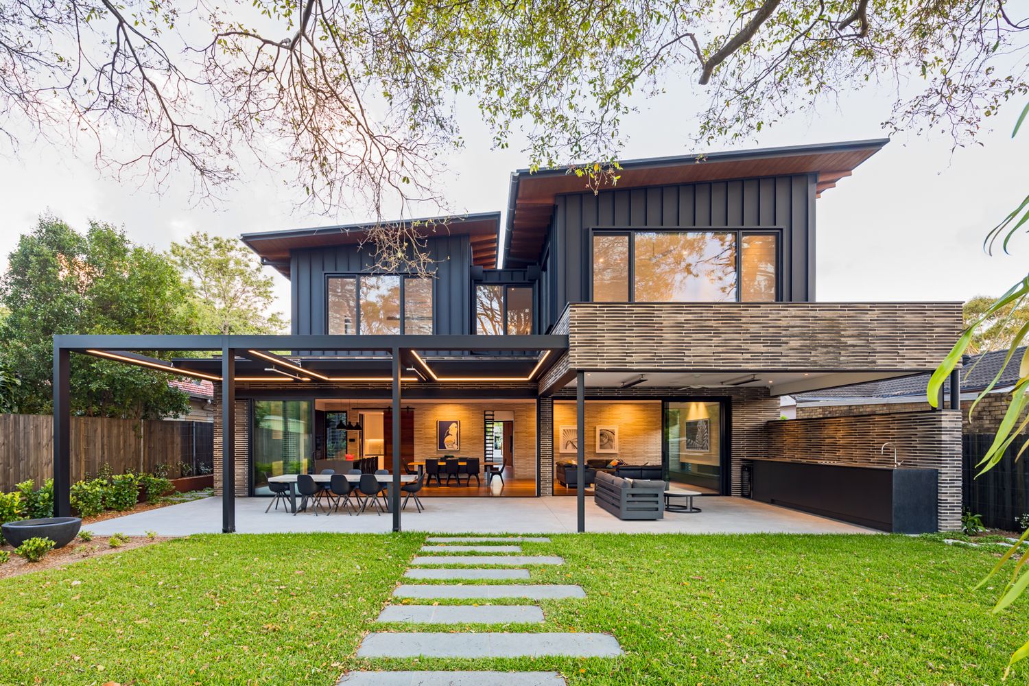 Contemporary dark timber and brick house with a large covered patio, stepping stone path and landscaped lawn under mature trees.