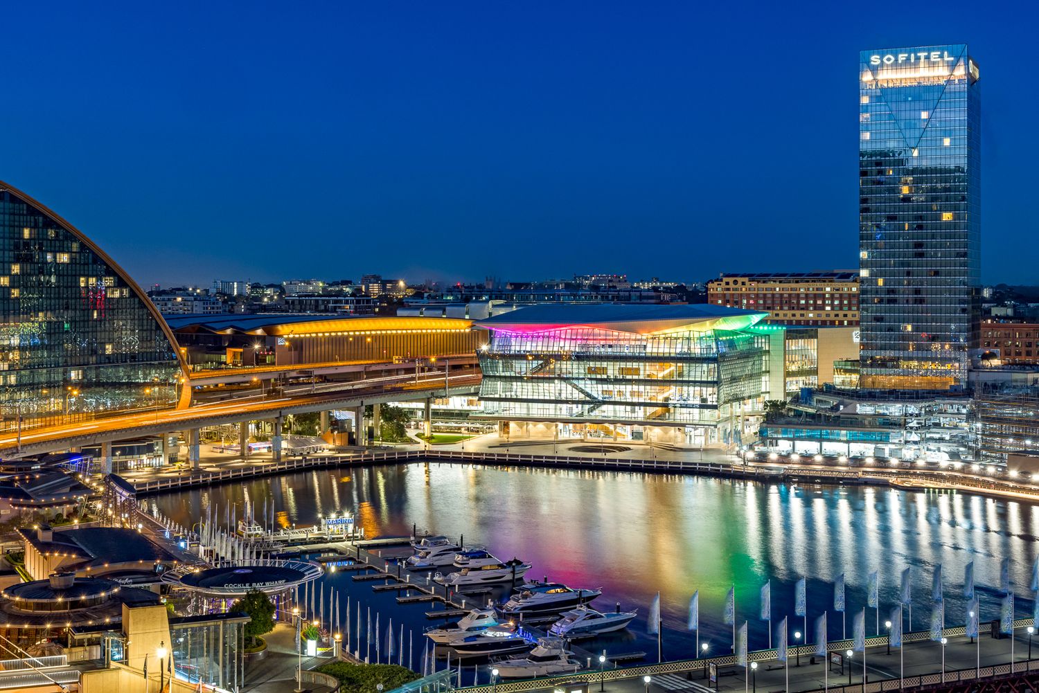 Sydney Darling Harbour at dusk with illuminated international convention centre, rainbow-lit facade, marina boats and a Sofitel hotel tower.