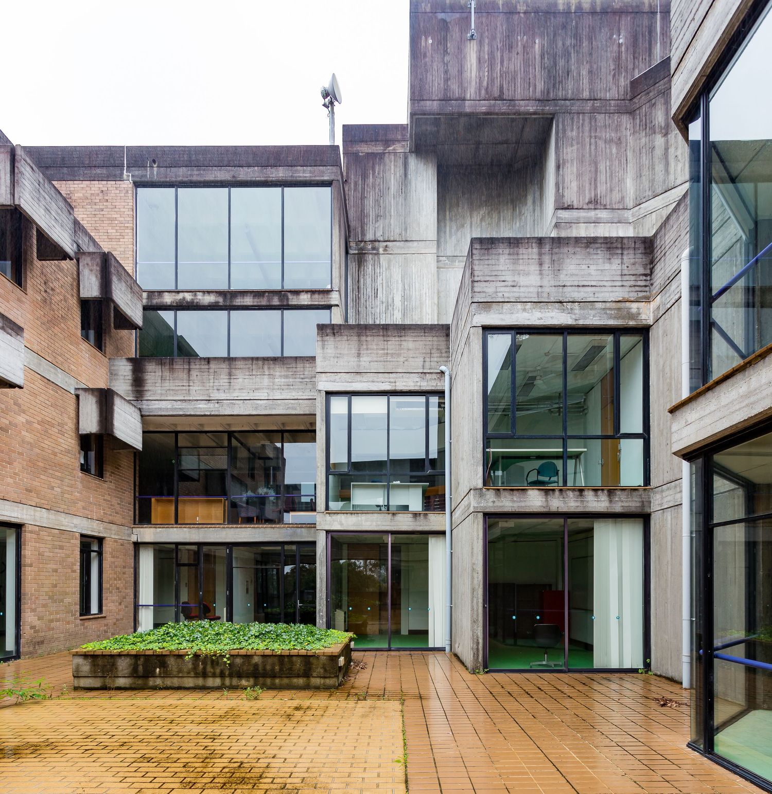 Brutalist concrete and brick building complex with large glass windows and a paved courtyard featuring a low planter of green plants.