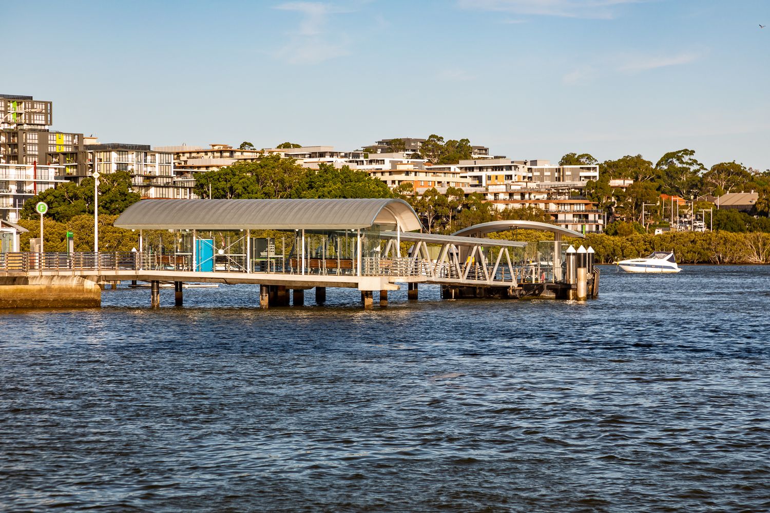 Covered ferry wharf on the harbour with waterfront apartments and green hillside trees rising in the background.