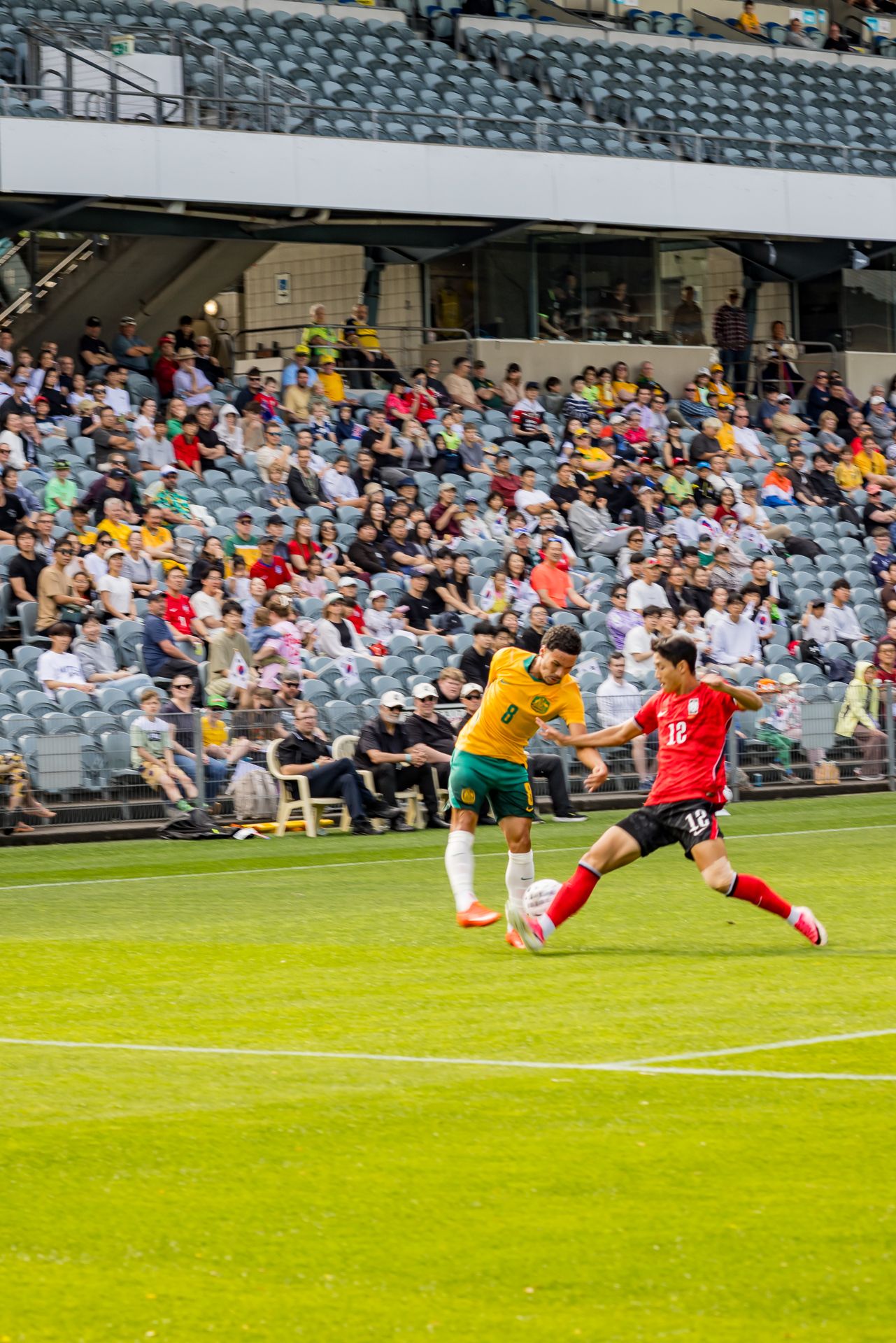 Australian player in green and gold challenges an opponent in red for the ball as spectators watch from the stadium stands.
