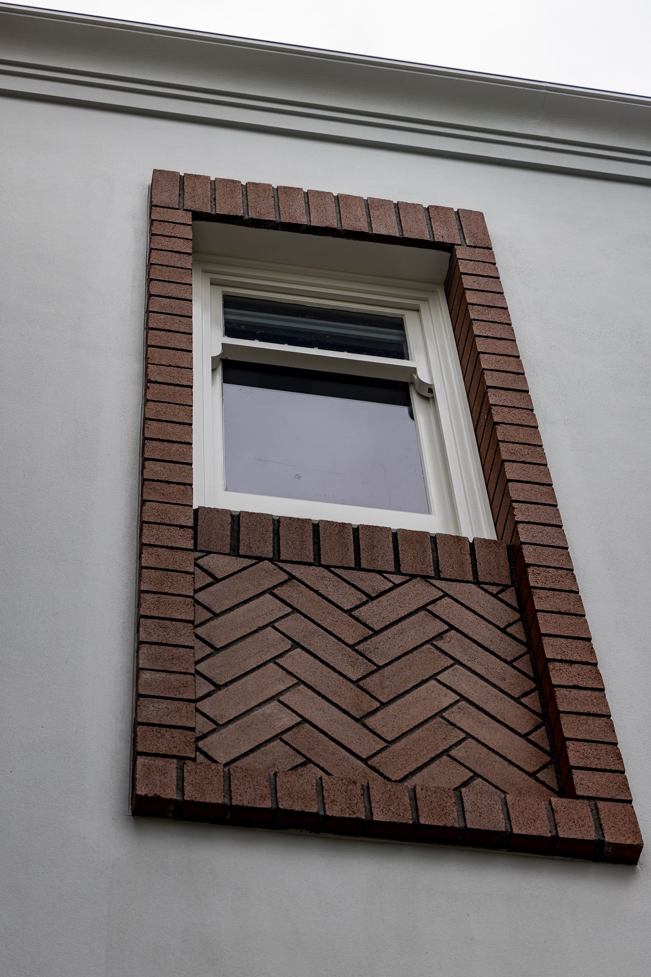 Upper-storey sash window framed by red brick surround with herringbone brick pattern on a grey rendered facade.
