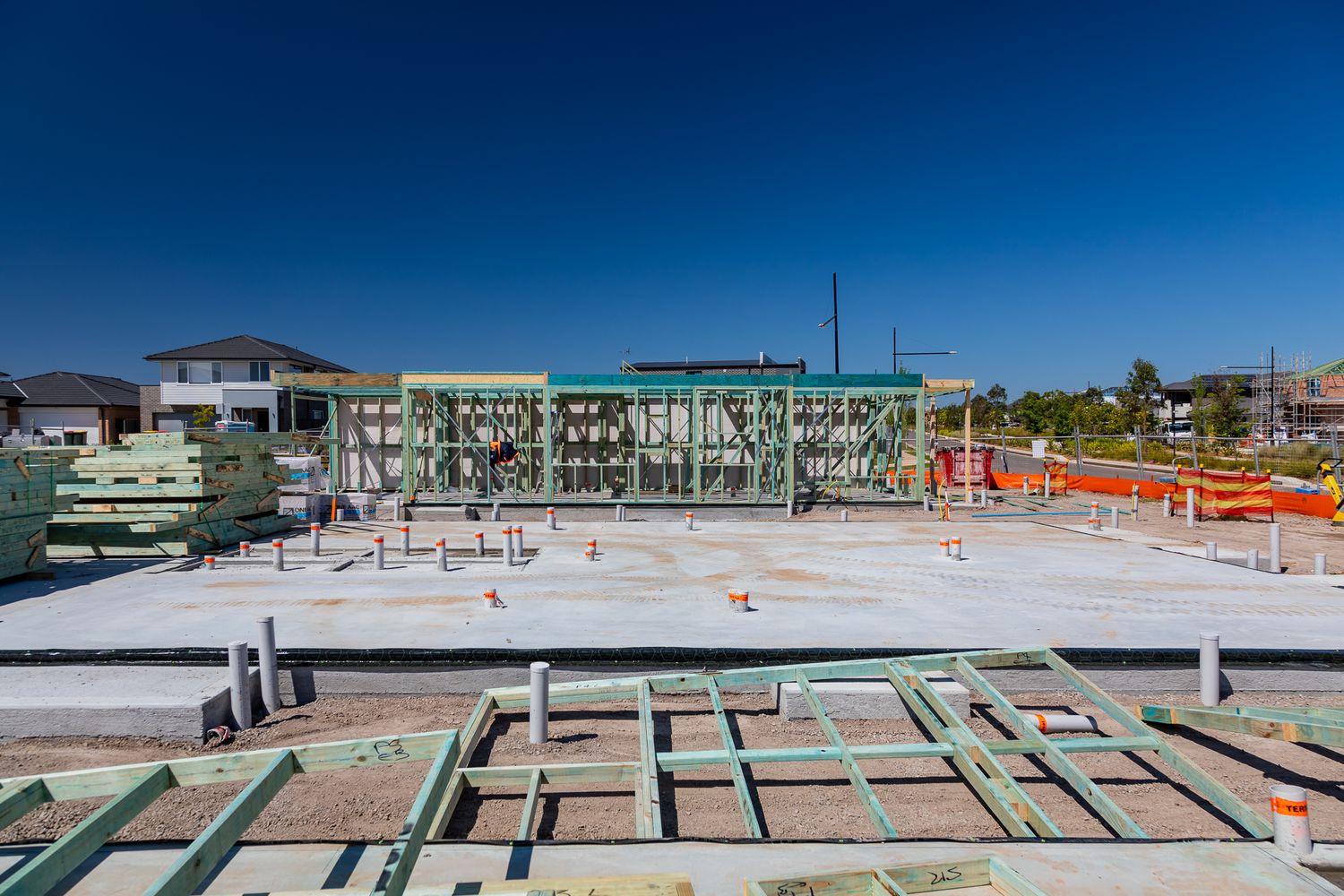 Concrete slab foundation with plumbing pipes and green steel framing rising at a residential building site under a clear blue sky.