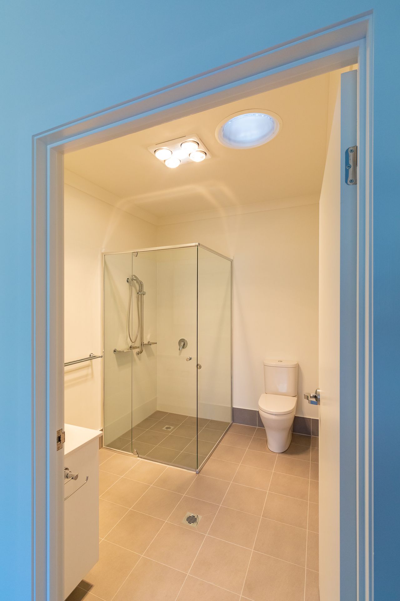 Modern bathroom with a frameless glass shower, tiled floor, white toilet, vanity and ceiling heat lamp seen through a blue doorway.