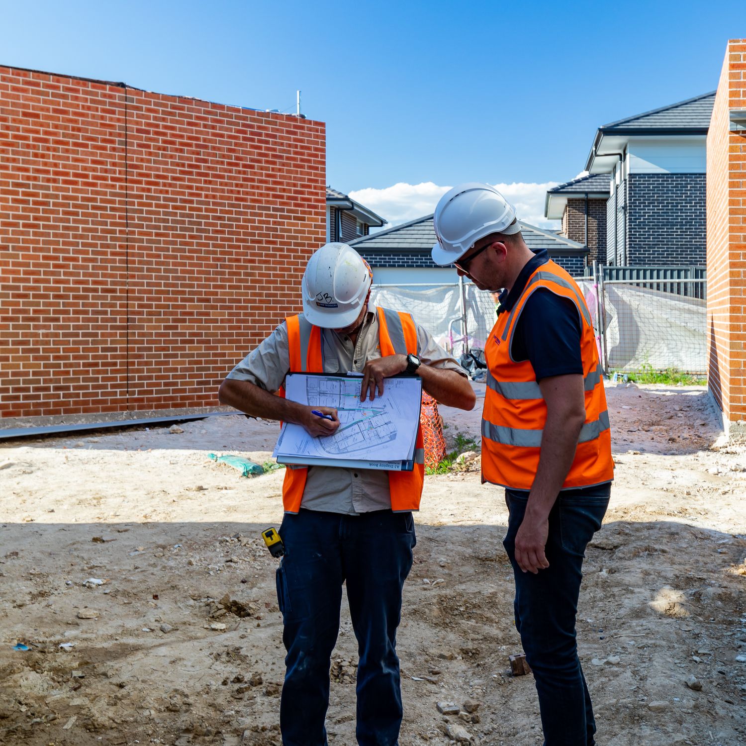 Two construction workers in hard hats and hi-vis vests review building plans on a site beside a new brick home.