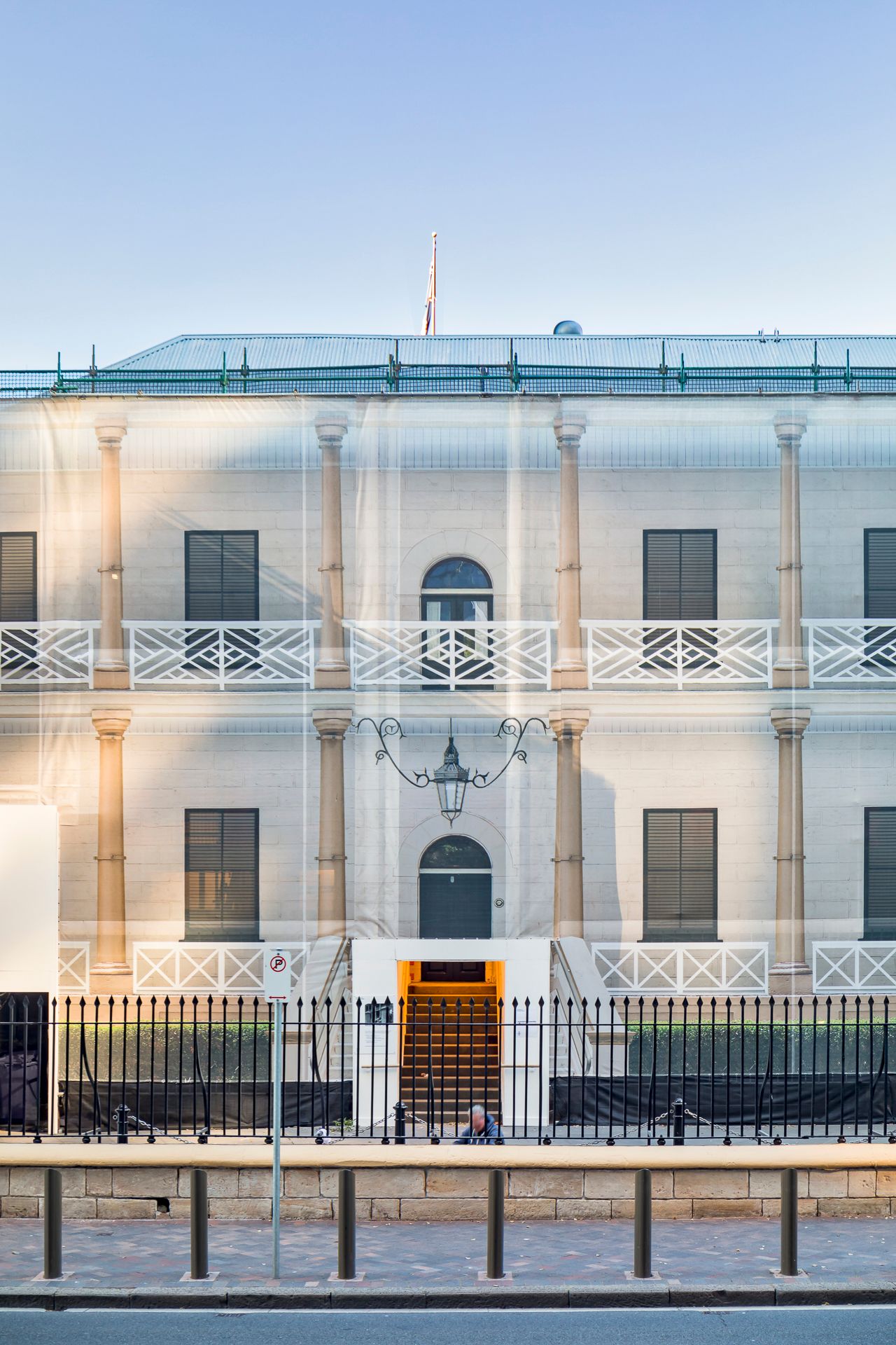 Neoclassical building facade wrapped in translucent protective sheeting with balconies, arched doorways and a black iron fence in front.
