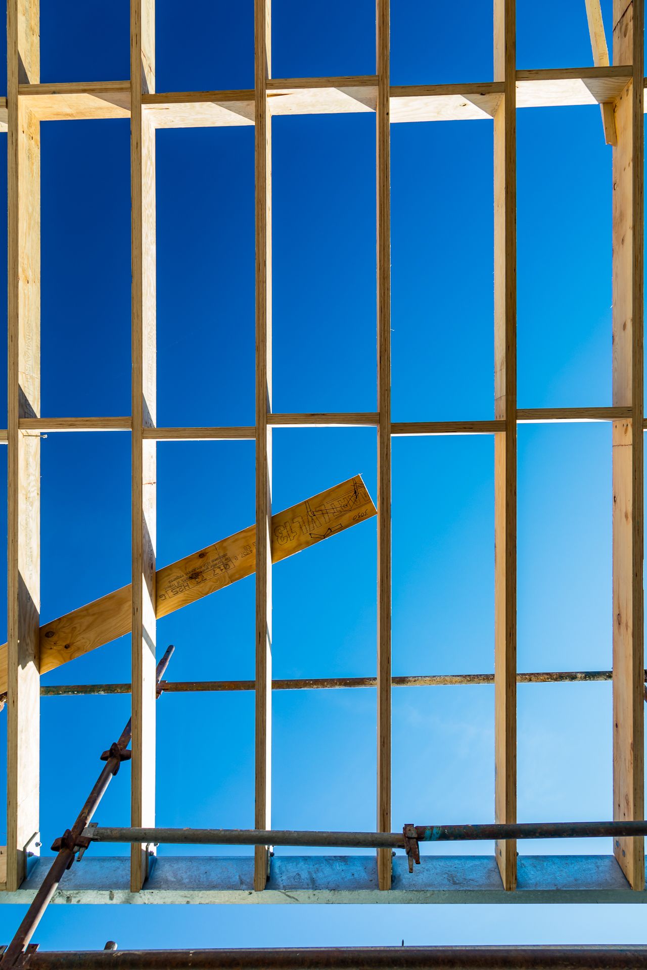 Vertical timber framing with a diagonal brace and scaffolding silhouetted against a clear blue sky on a construction site.