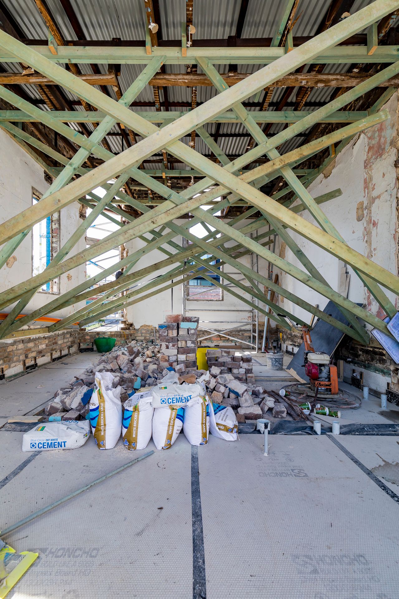 Attic renovation site with exposed timber roof trusses, corrugated roofing, bags of cement and a pile of bricks on the concrete floor.