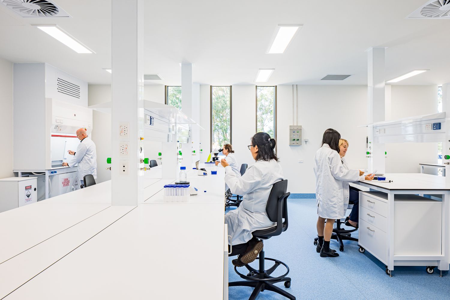 Scientists in white coats conduct experiments at benches in a bright, open laboratory with windows overlooking greenery.
