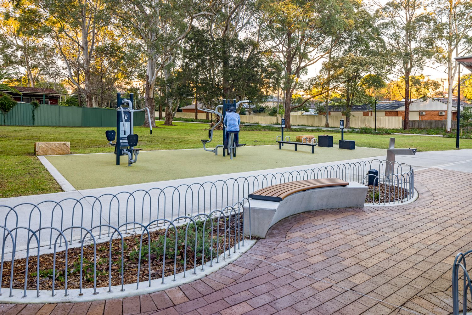 Outdoor fitness equipment on a rubberised surface beside a curved brick seating wall and paved path in a leafy suburban park.