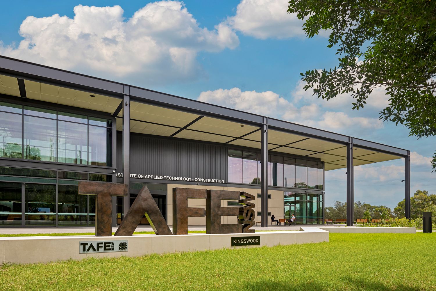 Entrance to a modern TAFE NSW applied trades building with large timber lettering, glass facade, and green lawn beneath a cloudy blue sky.