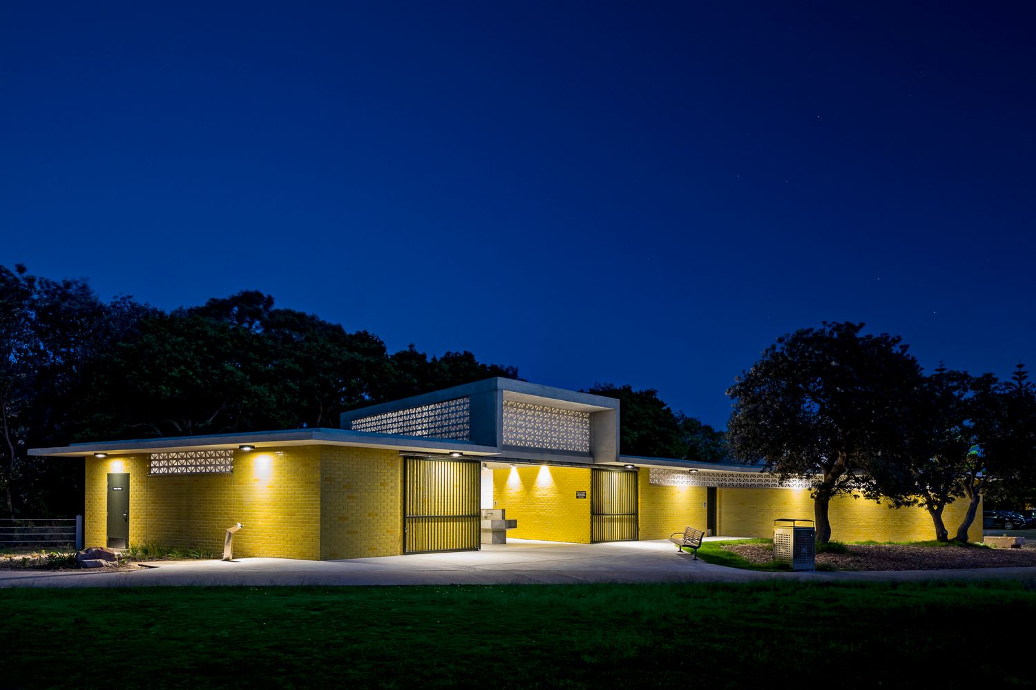 Modern yellow brick public restroom building glowing at dusk with warm lighting, surrounded by trees and a lawn.