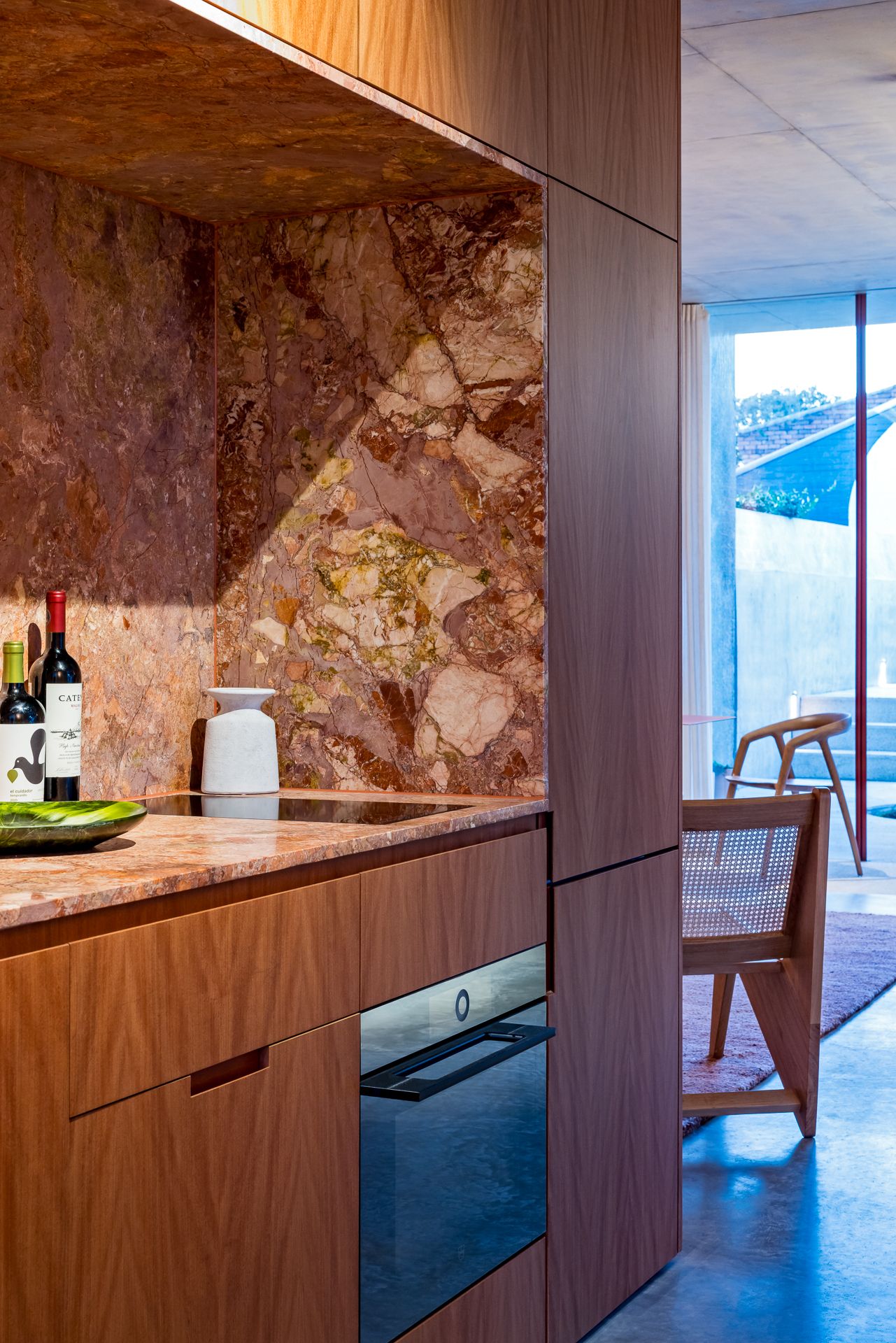 Modern kitchen with warm walnut cabinetry, a built-in oven, and a red marble splashback beside a window and woven wooden chair.