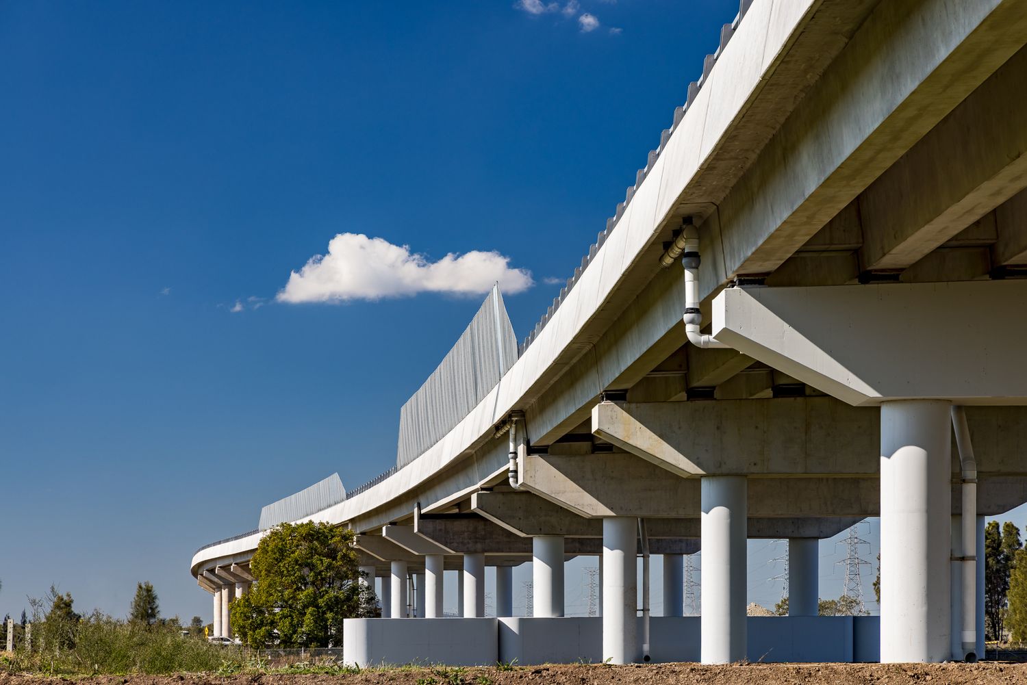Upward view of curved concrete highway bridge with rounded pillars against a deep blue sky and white cloud.