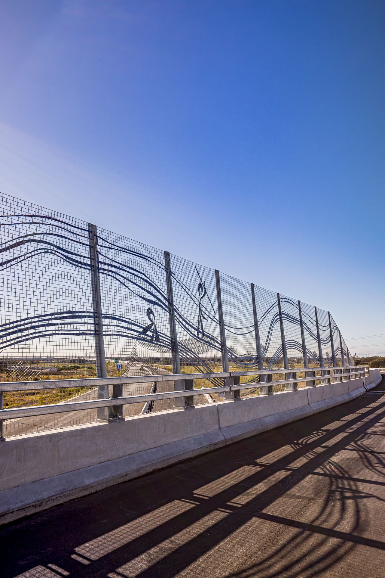 Bridge safety barrier with flowing wave line artwork and figure silhouettes in steel mesh, overlooking a highway under blue sky.