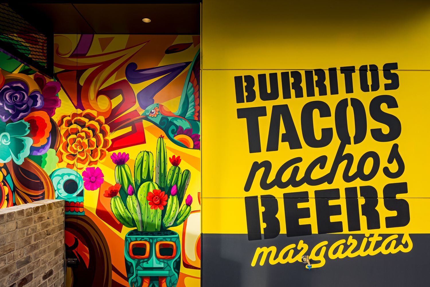 Bold black lettering on a yellow wall advertising burritos, tacos, nachos, beers and margaritas beside a colorful cactus mural.